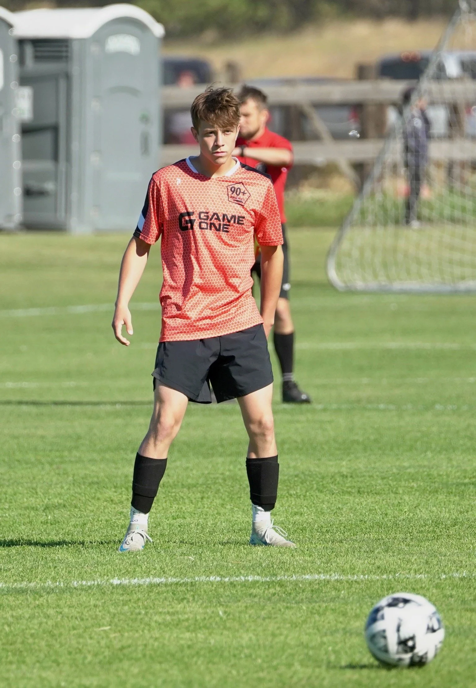 A young male soccer player standing on a soccer field, wearing a red jersey with black shorts, black socks, and white cleats, preparing to kick a soccer ball.