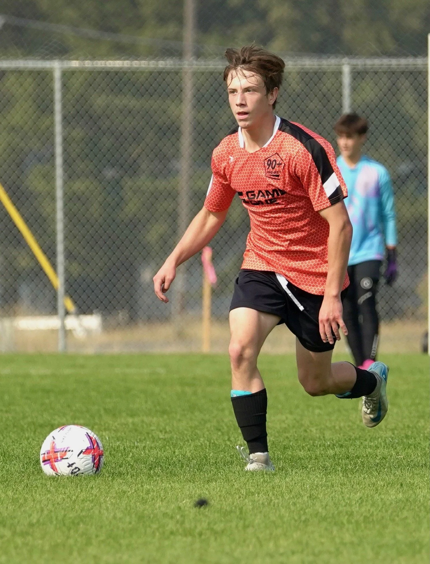 Young male soccer player in orange jersey and black shorts dribbling a white soccer ball with red and purple accents on a grassy field.