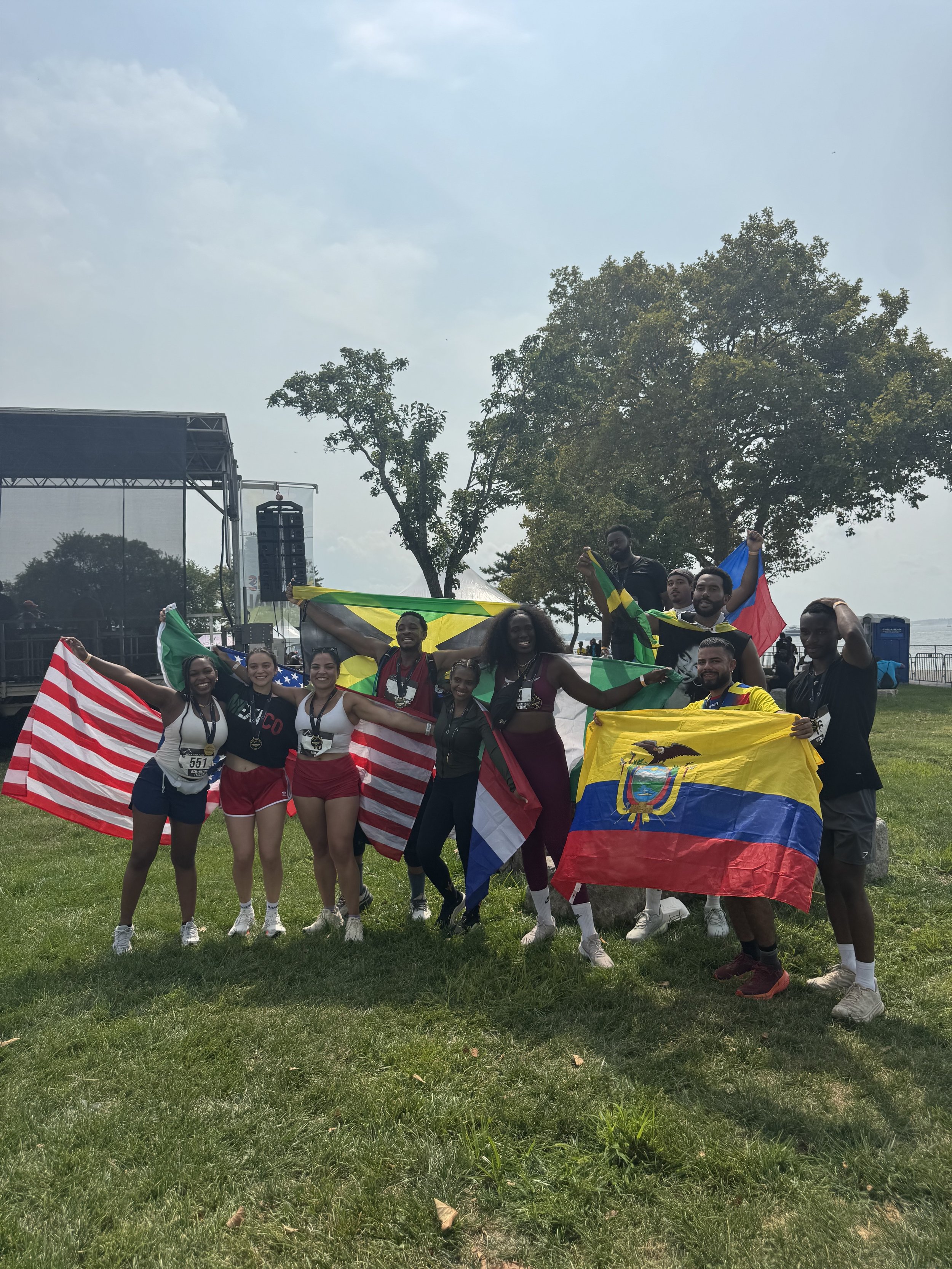 A group of diverse athletes celebrating with flags from different countries, standing on grass with trees and a stage in the background.