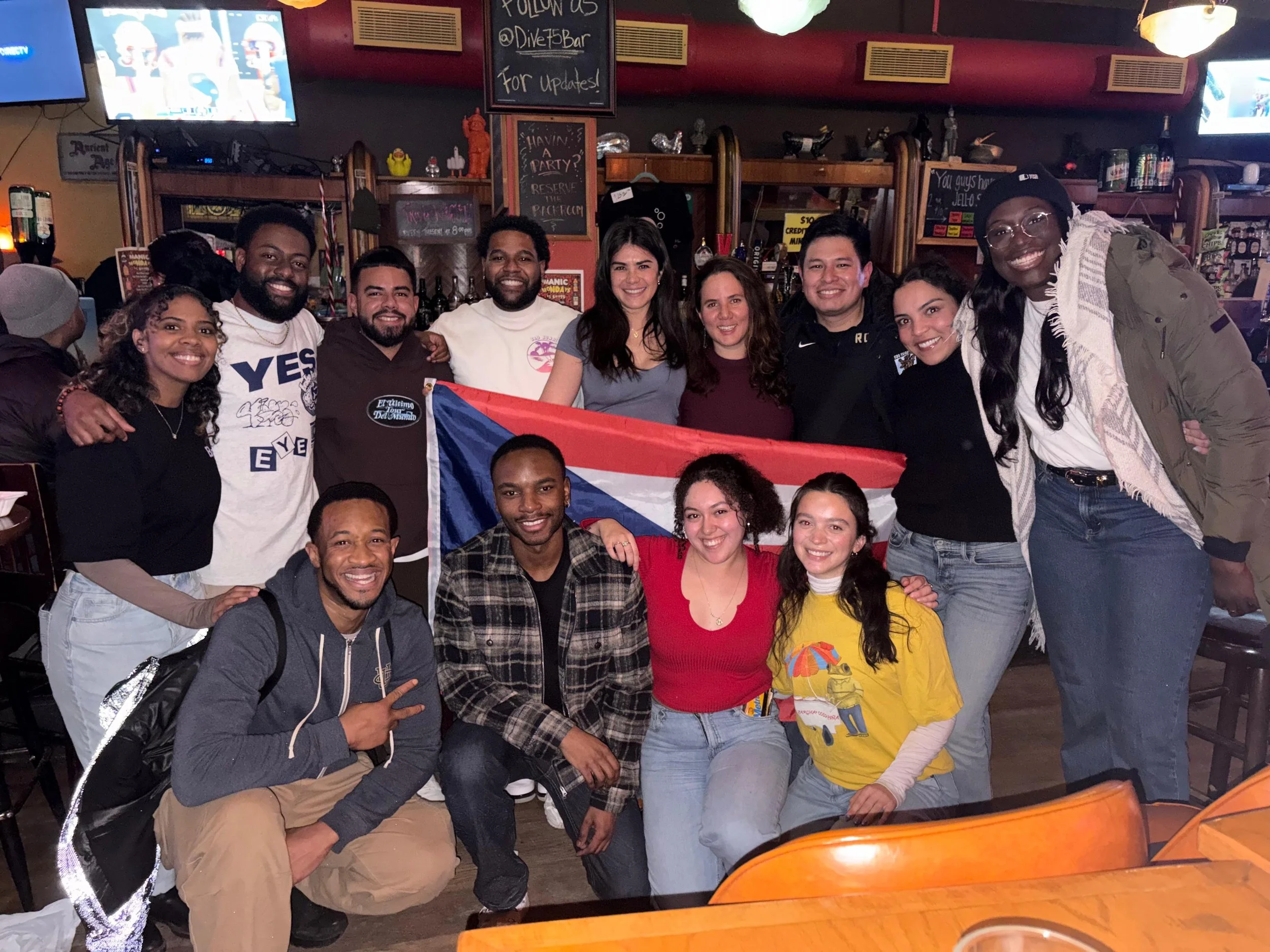A group of people in a bar holding a Puerto Rican flag, smiling at the camera.