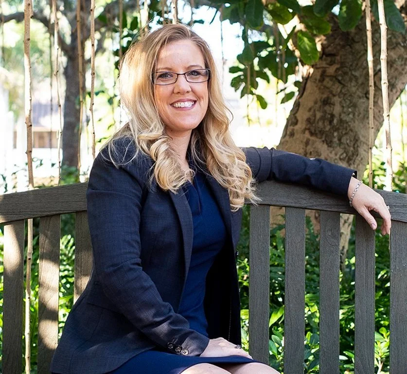 Woman sitting on a bench with trees in the background, wearing glasses, a dark blazer, and a blue dress. She is smiling and has long, wavy blonde hair.