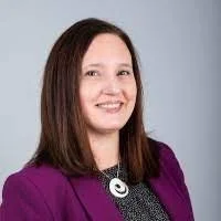 Portrait of a smiling woman with long brown hair wearing a purple blazer and a black patterned top with a necklace, against a plain background.