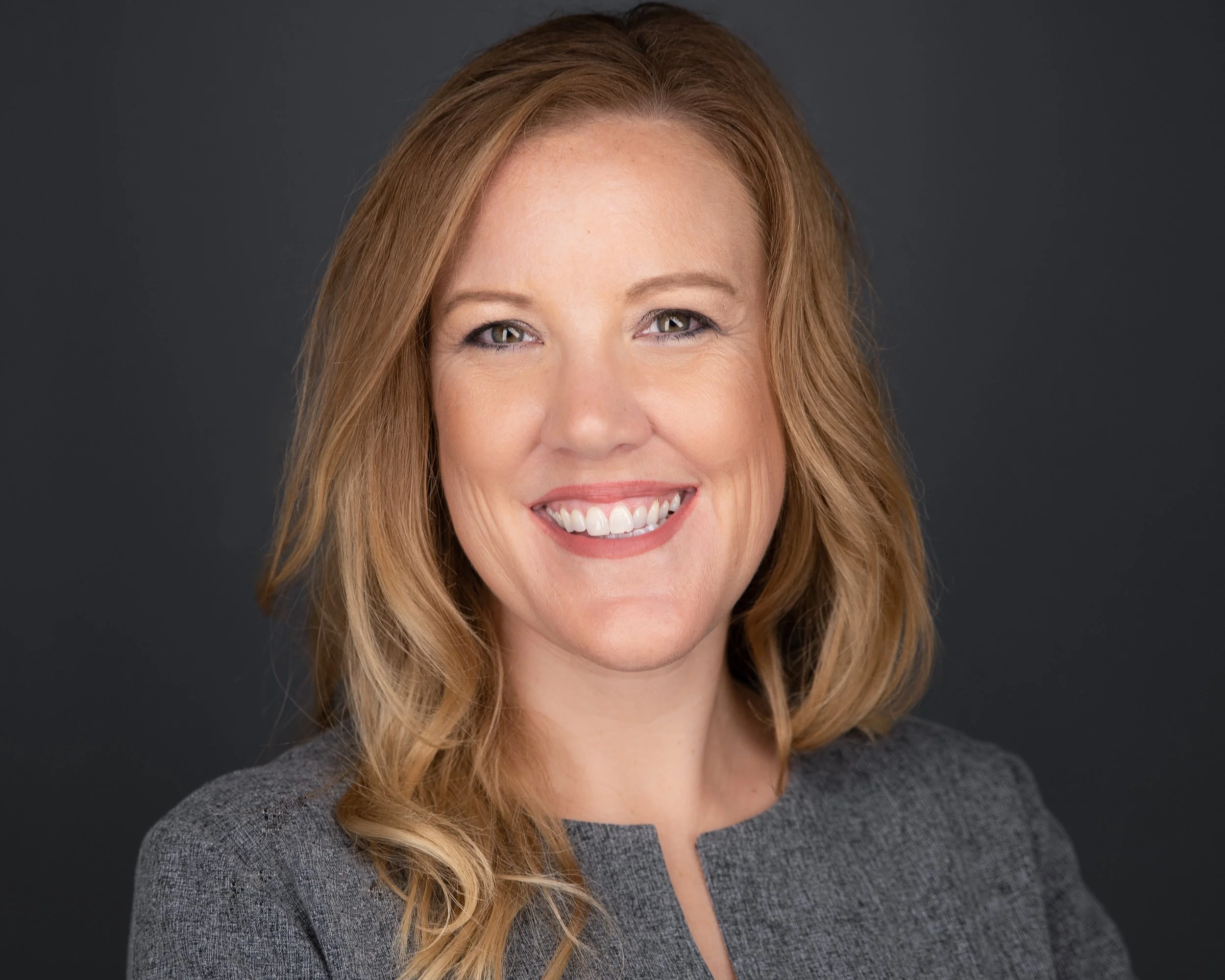 Smiling woman with wavy blonde hair in a gray blazer, dark background.