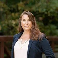 Woman standing outdoors in business attire, posing for a professional portrait with greenery in the background.