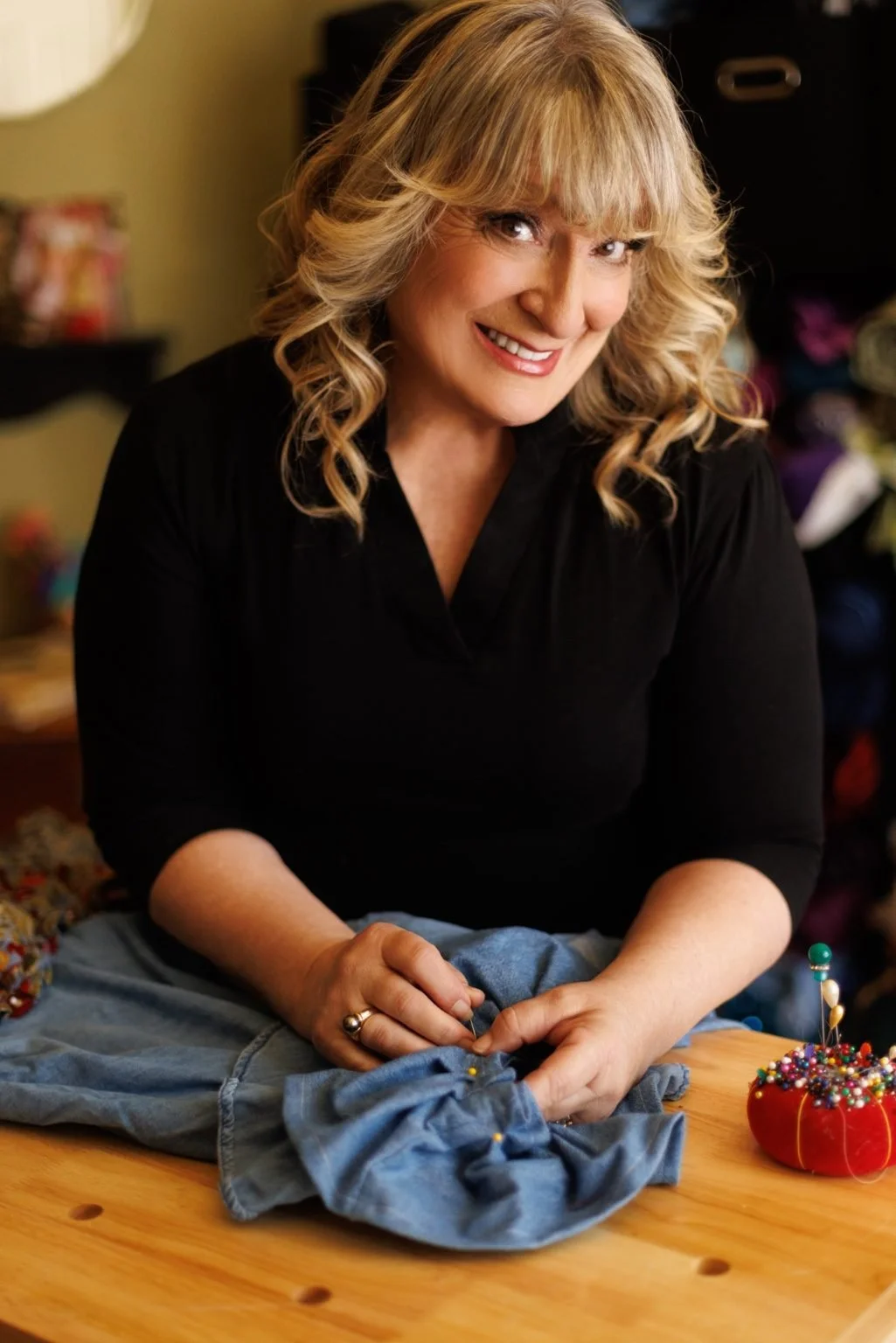 Portrait of Sandy Beers, Costumer for EnKore Theatre Company, smiling while sewing fabric at a worktable with sewing pins and costume materials nearby.