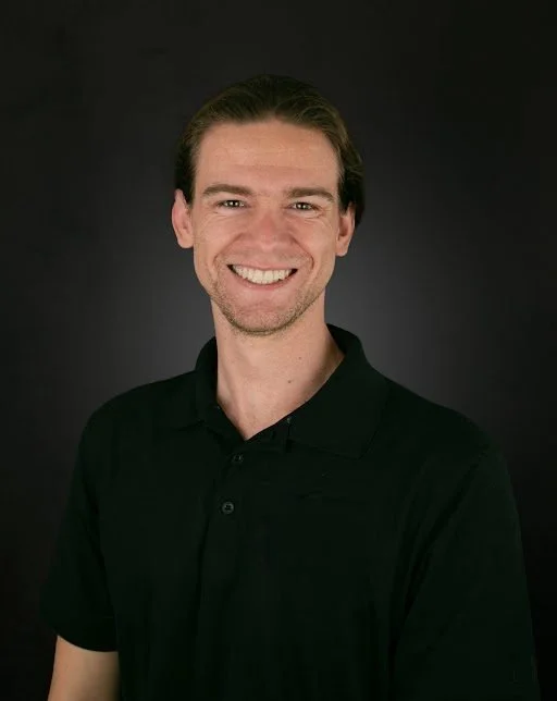 Portrait of Alex May, Technical Director and Co-Founder of EnKore Theatre Company, smiling while wearing a black polo shirt against a dark background.