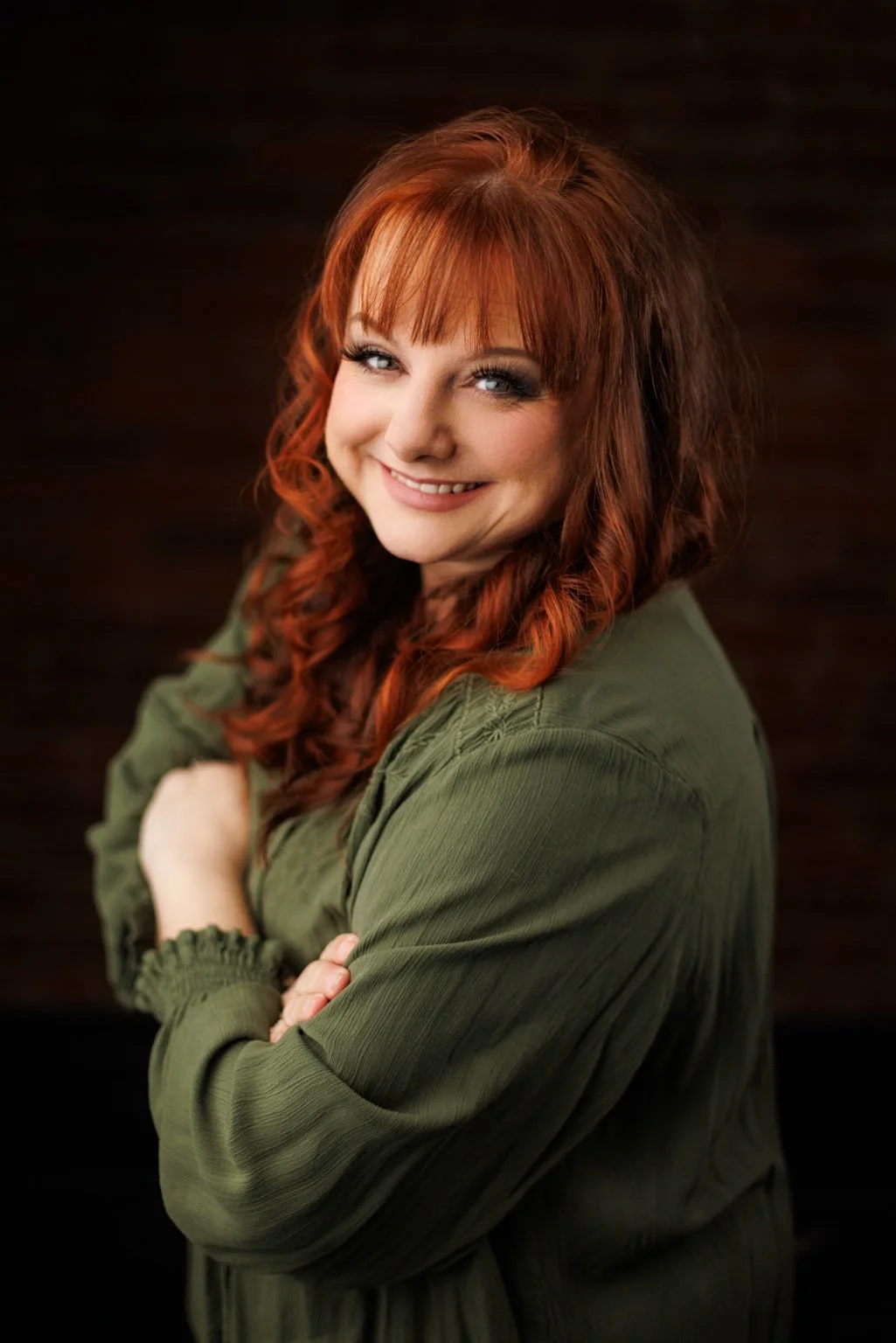 Portrait of Kelly Lynn Jordan, Executive Director and Co-Founder of EnKore Theatre Company, smiling while wearing a green blouse against a dark background.