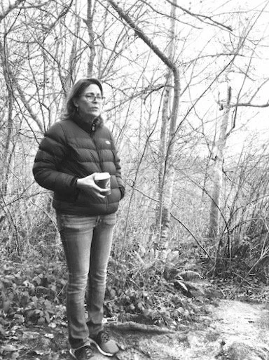 A woman standing outdoors near a creek in a wooded area during winter, holding a cup.