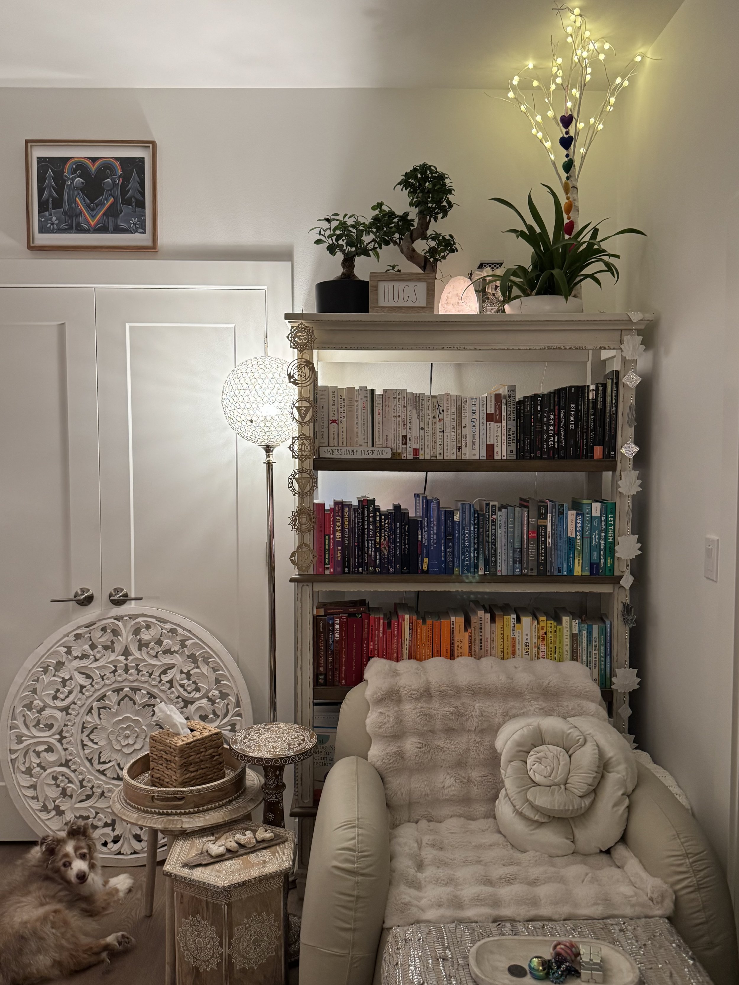 Cozy living room corner with a white bookshelf filled with books organized by color, a plush cream-colored armchair with a fur throw and a spiral pillow, a small side table with a decorative tray and some stones, a dog lying on the floor, a standing lamp, a potted plant, and wall art with love-themed illustration.