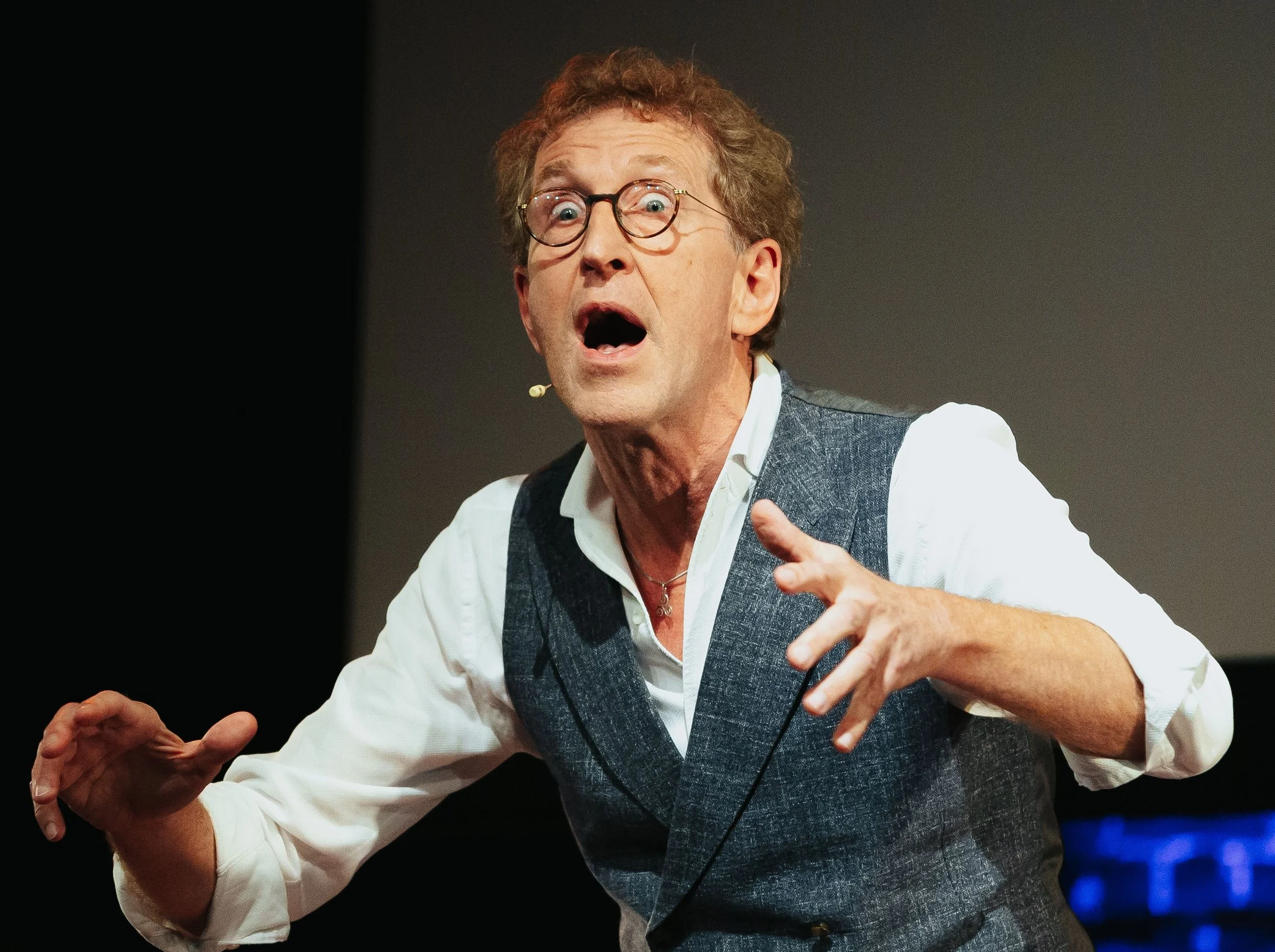 Homme en costume gris avec lunettes, parlant passionnément lors d'une présentation ou spectacle.