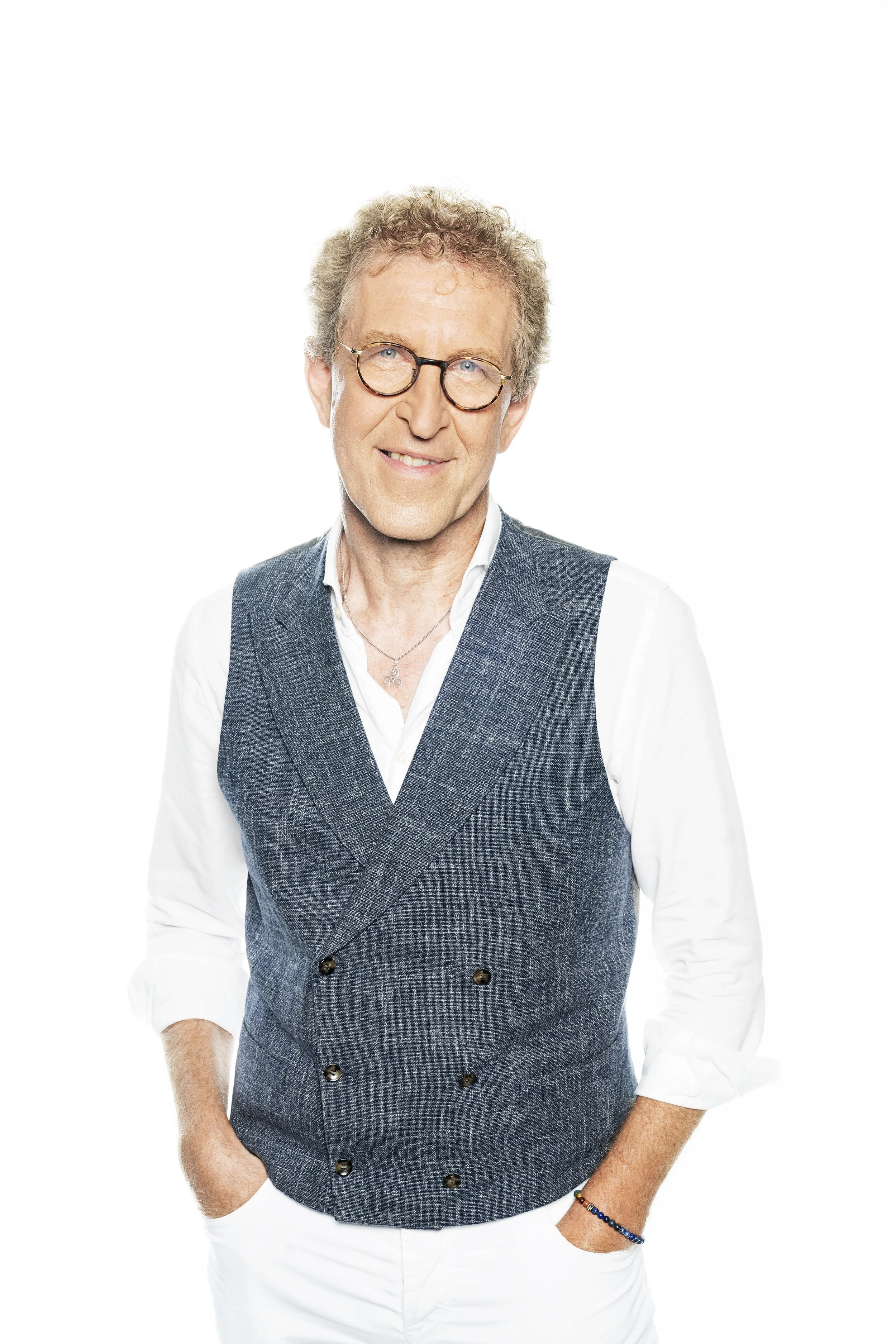 Homme âgé souriant portant une veste en tweed, une chemise blanche et des lunettes, avec un fond blanc.