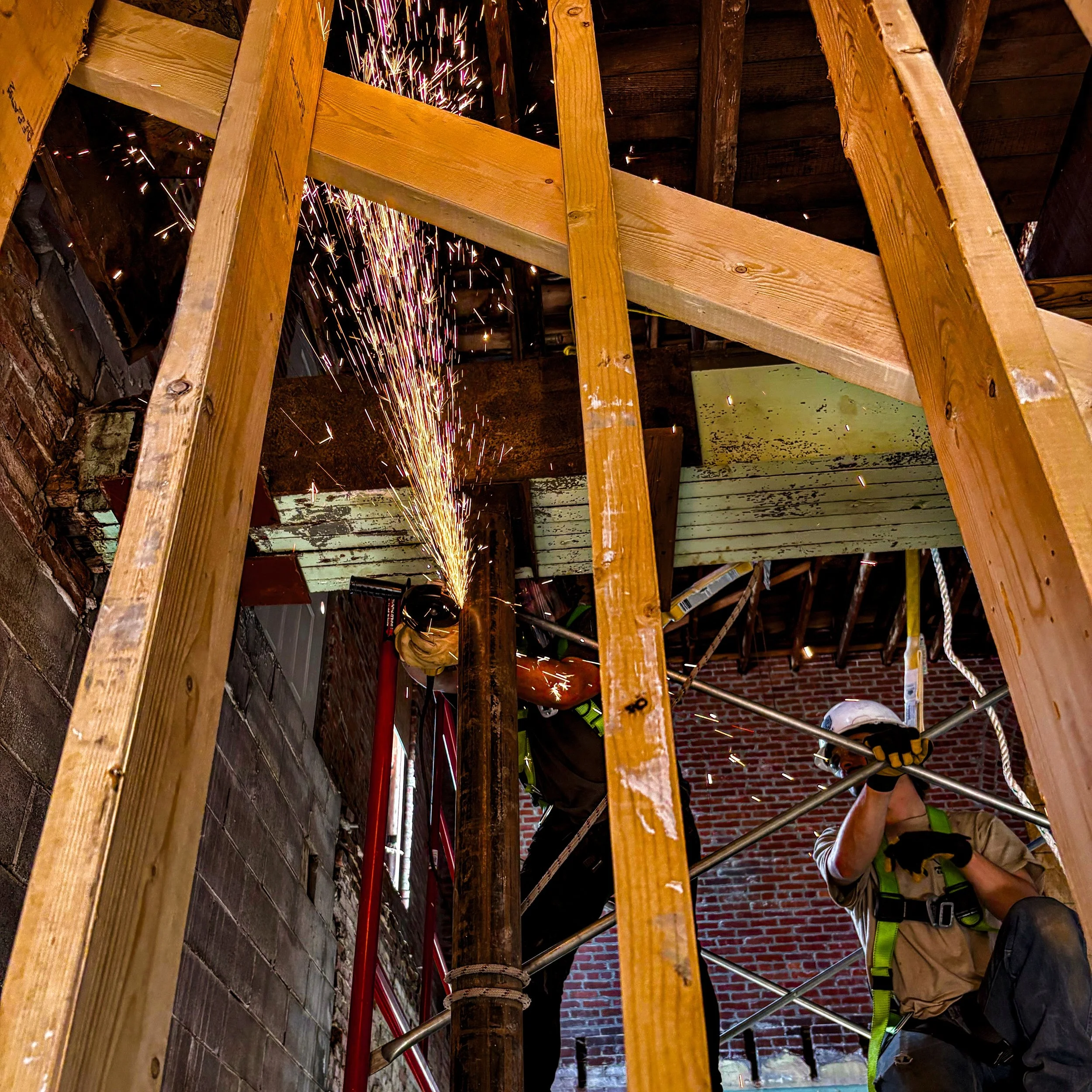 A construction worker wearing a helmet and safety harness uses a welding tool on an old steel pipe inside a building, with sparks flying from the grinding process.