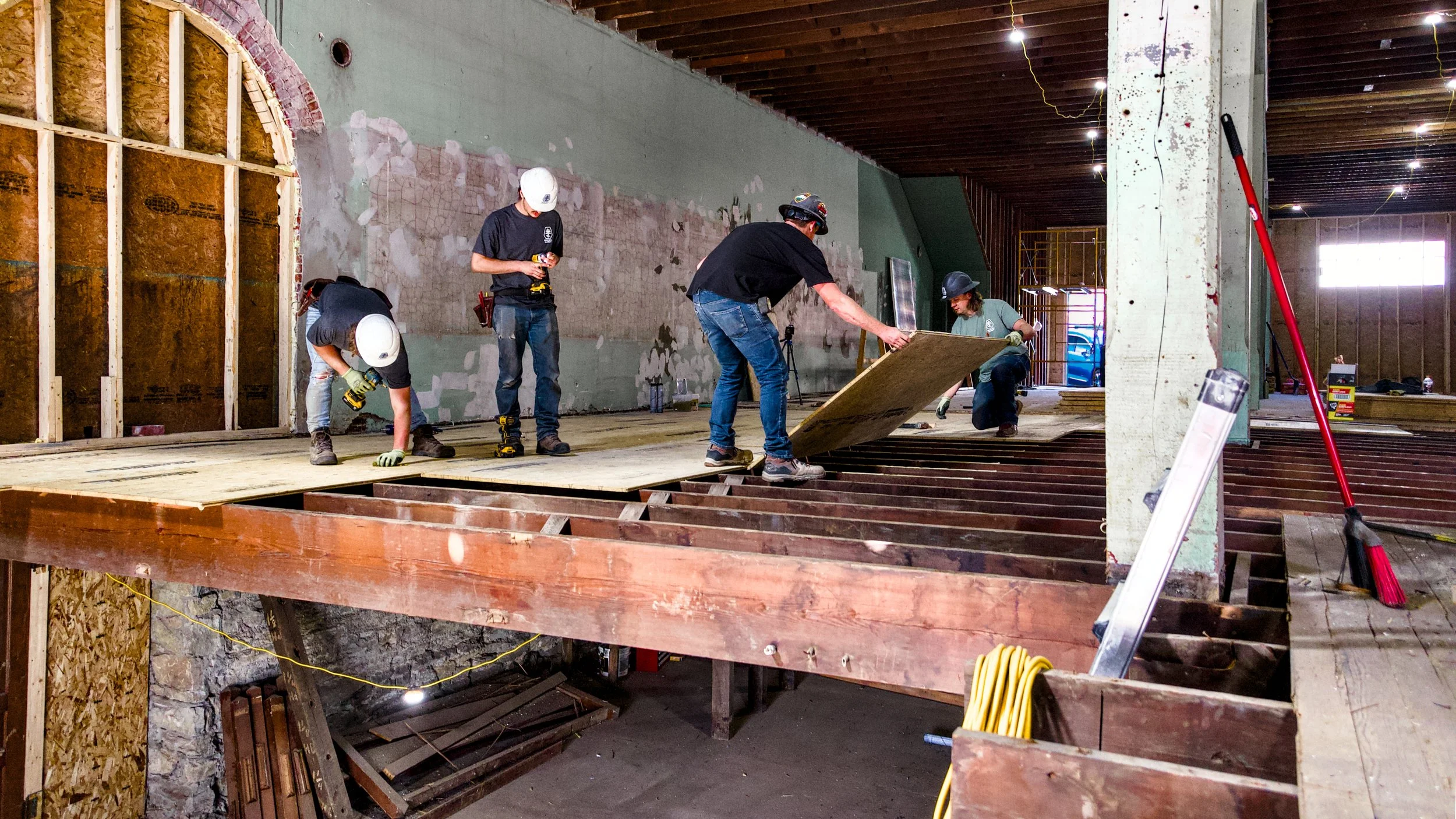 Construction workers installing a sub-floor on a building's upper level, with framing and unfinished walls visible.