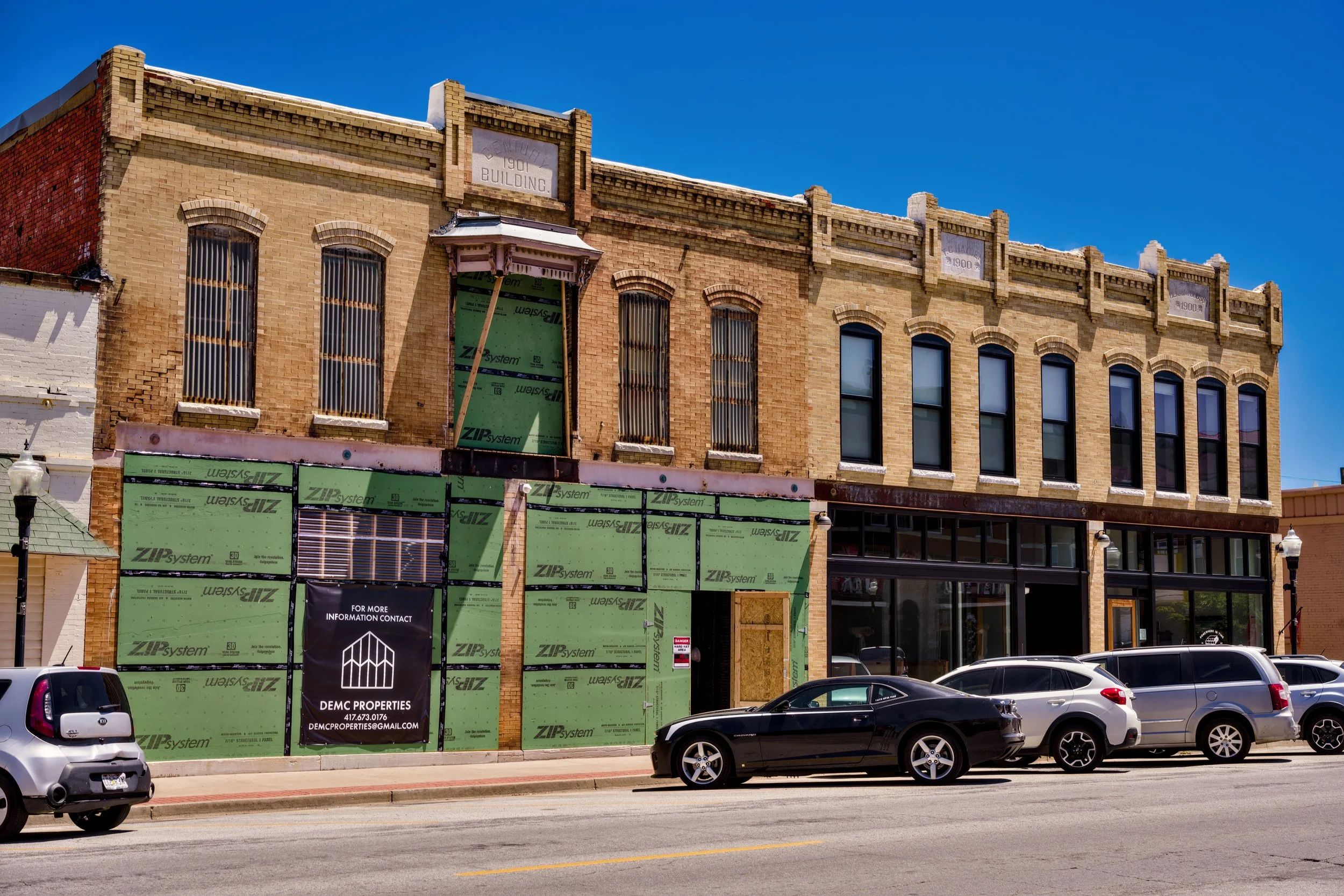 A multi-story brick building under construction or renovation with green sheathing on the lower part, and with unfinished upper floors. Several parked cars are in front of the building on a city street, with a clear blue sky overhead.