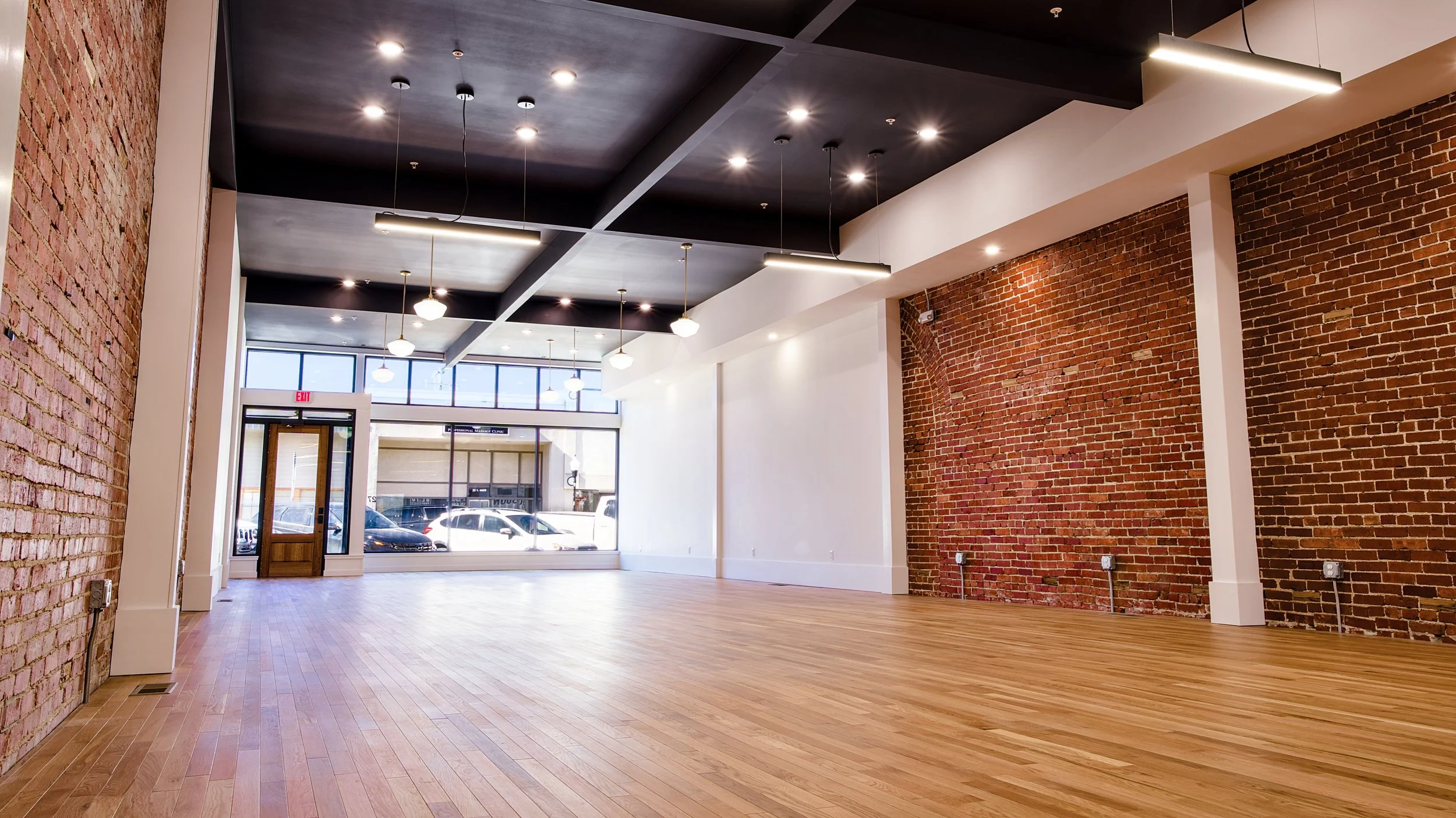 Empty interior space with hardwood floors, exposed brick walls, black ceiling with recessed lights, hanging light fixtures, large front windows, and a glass door exit.