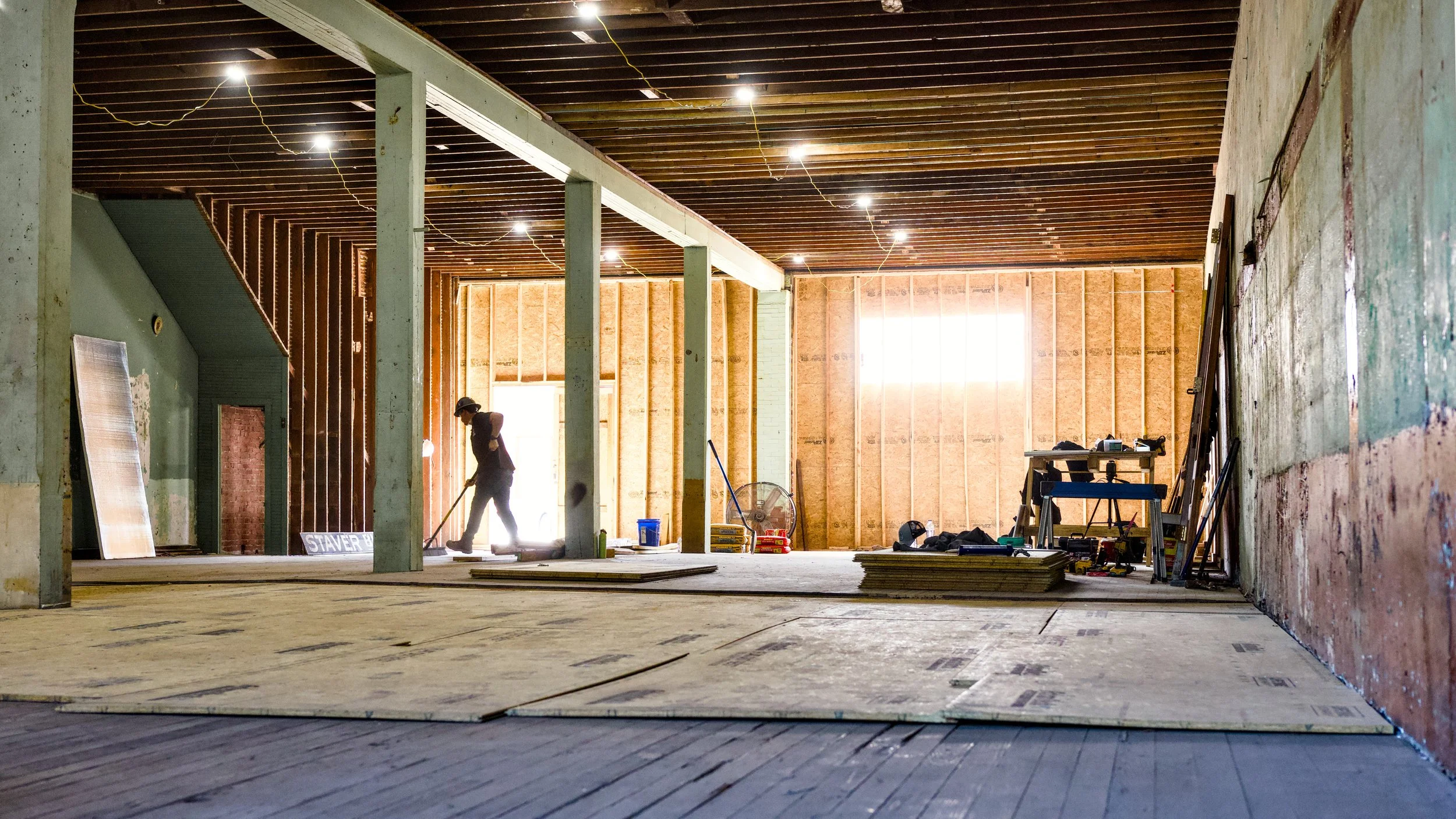 Interior of a house under construction with exposed wooden walls and ceiling, construction tools, and a worker sweeping the floor.
