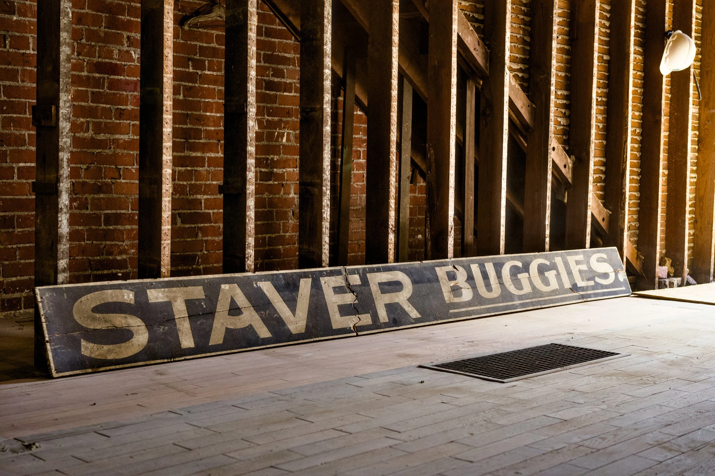 An indoor scene with a brick wall and wooden floor, featuring a weathered, cracked sign that reads "STAYER BUGGIES" and a partially visible wooden staircase. The lighting is warm and dim, and there is a wall-mounted lamp on the right side.