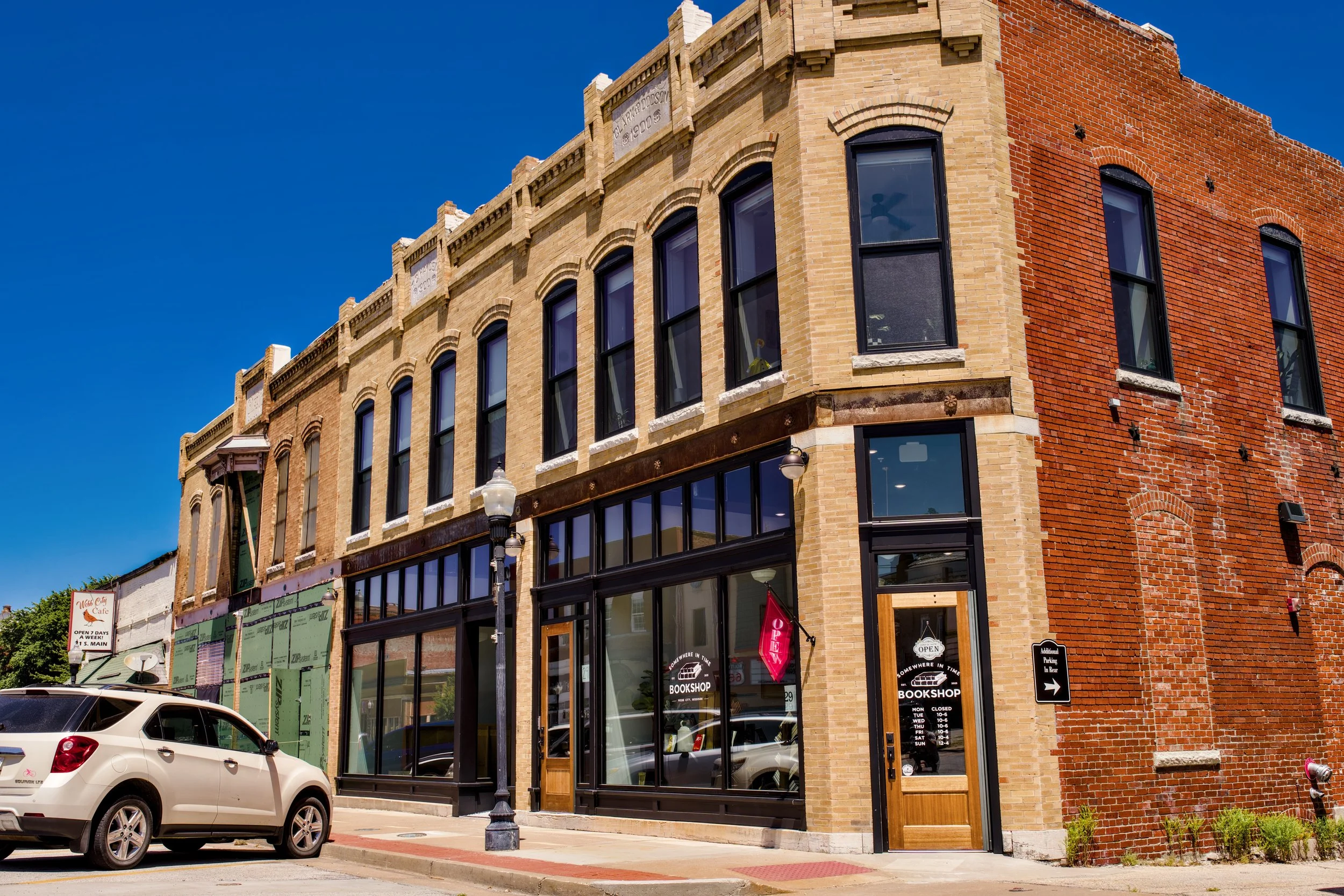 The corner of a historic brick building with large glass storefront windows and a wooden entrance door. The building has decorative brickwork and a sign indicating it is a bookstore. A small white car is parked in front, and there are street lamps on the sidewalk. The sky is clear and blue.