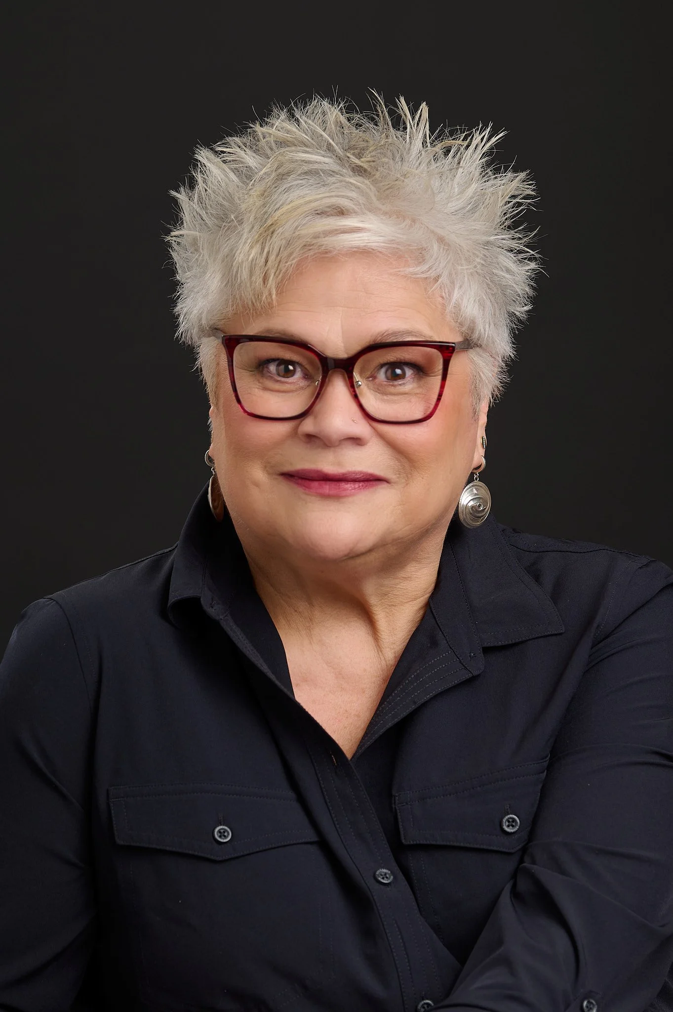 A woman with short, spiky gray hair, red glasses, wearing earrings and a black button-up shirt, smiling against a black background.