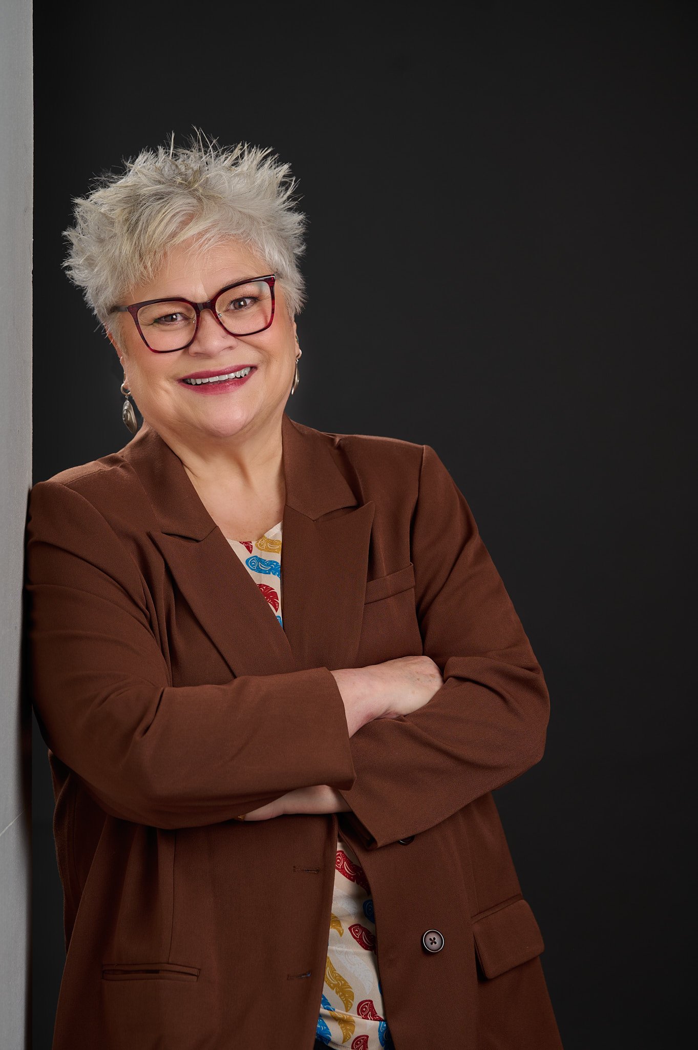 A smiling senior woman with short, curly gray hair and glasses, wearing a brown blazer and patterned shirt, standing with arms crossed against a black background.