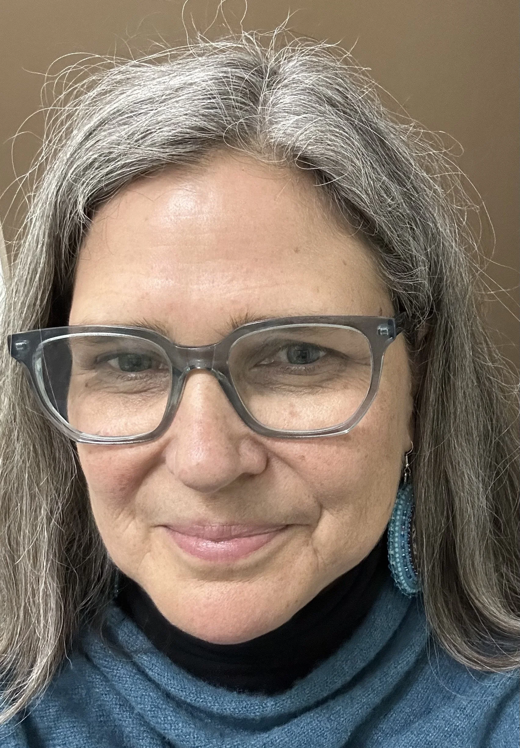 Close-up of a middle-aged woman with gray hair and glasses, smiling, wearing a black turtleneck and blue sweater.