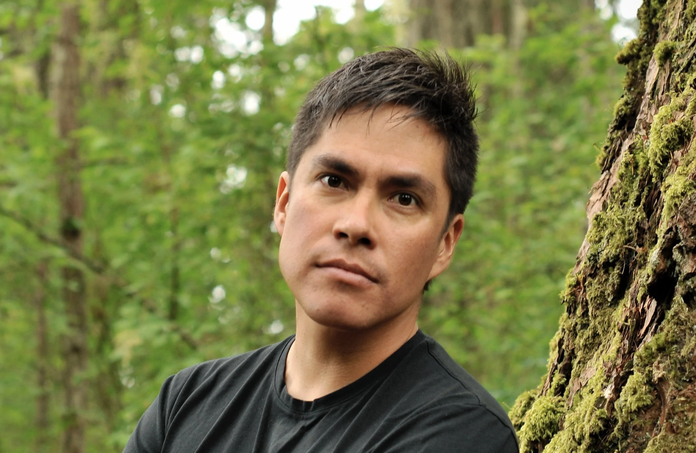 A man with short dark hair and fair skin, wearing a black T-shirt, outdoors in a forest, leaning against a tree covered in moss, with a blurred green leafy background.