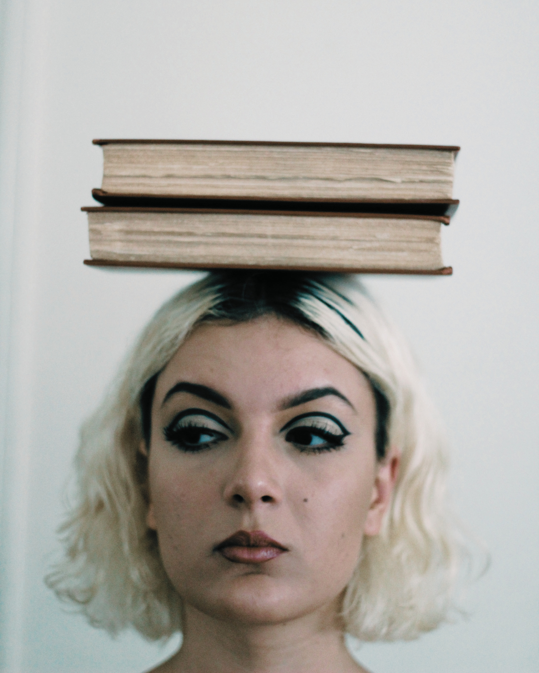 A woman with platinum blonde hair and bold makeup balancing two thick books on her head.