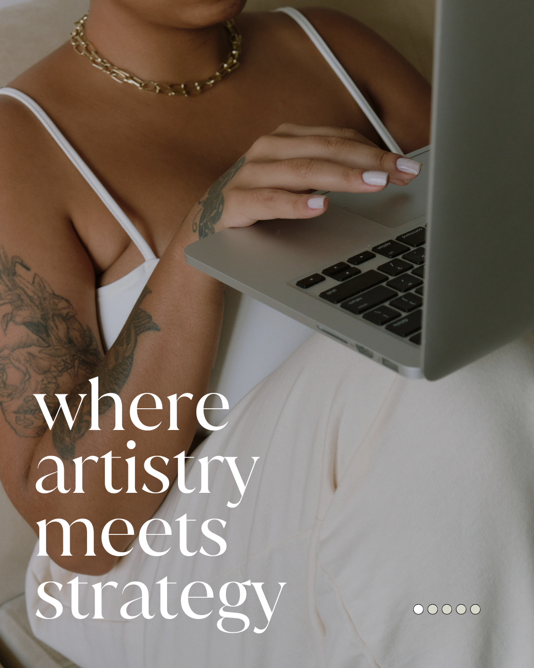 A woman with tattoos on her arm wearing a white top and gold necklace, using a laptop computer.