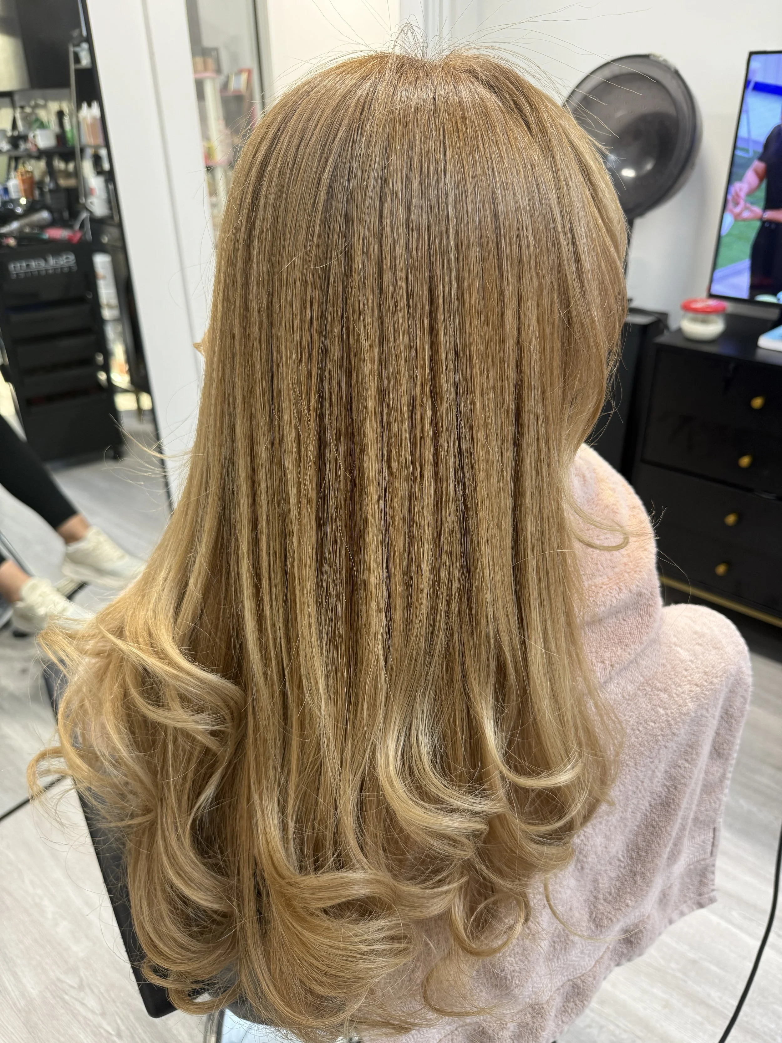 Back view of a woman with long, wavy, golden blonde hair in a salon setting.