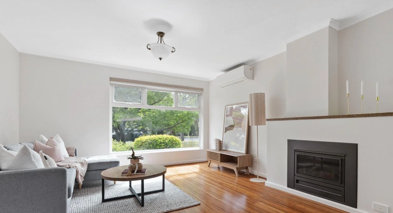Bright living room with large window, gray sofa, coffee table, fireplace, hardwood floors, and minimalist decor.