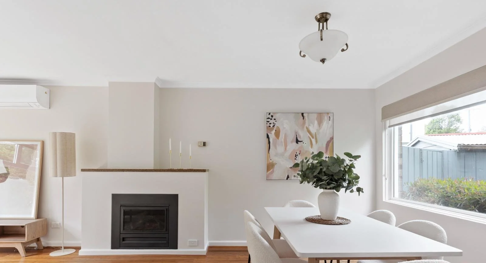 Bright living room with a white dining table, beige chairs, and a large window with a view of outside greenery. Art on the wall, fireplace, potted plant centerpiece, floor lamp, and ceiling light fixture.