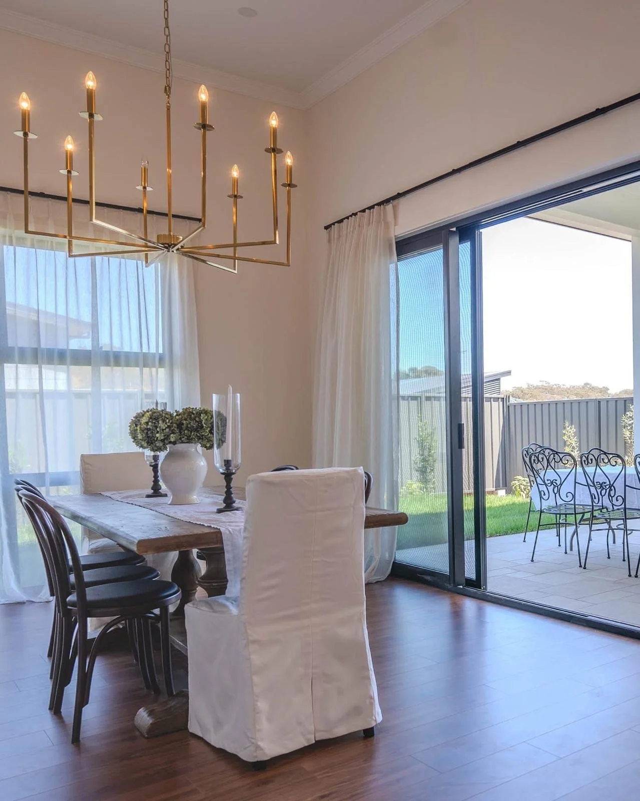 Dining room with a wooden table, white and black chairs, a white vase with flowers, and a modern chandelier, with sliding glass doors leading to an outdoor patio with metal chairs and a fence in the background.