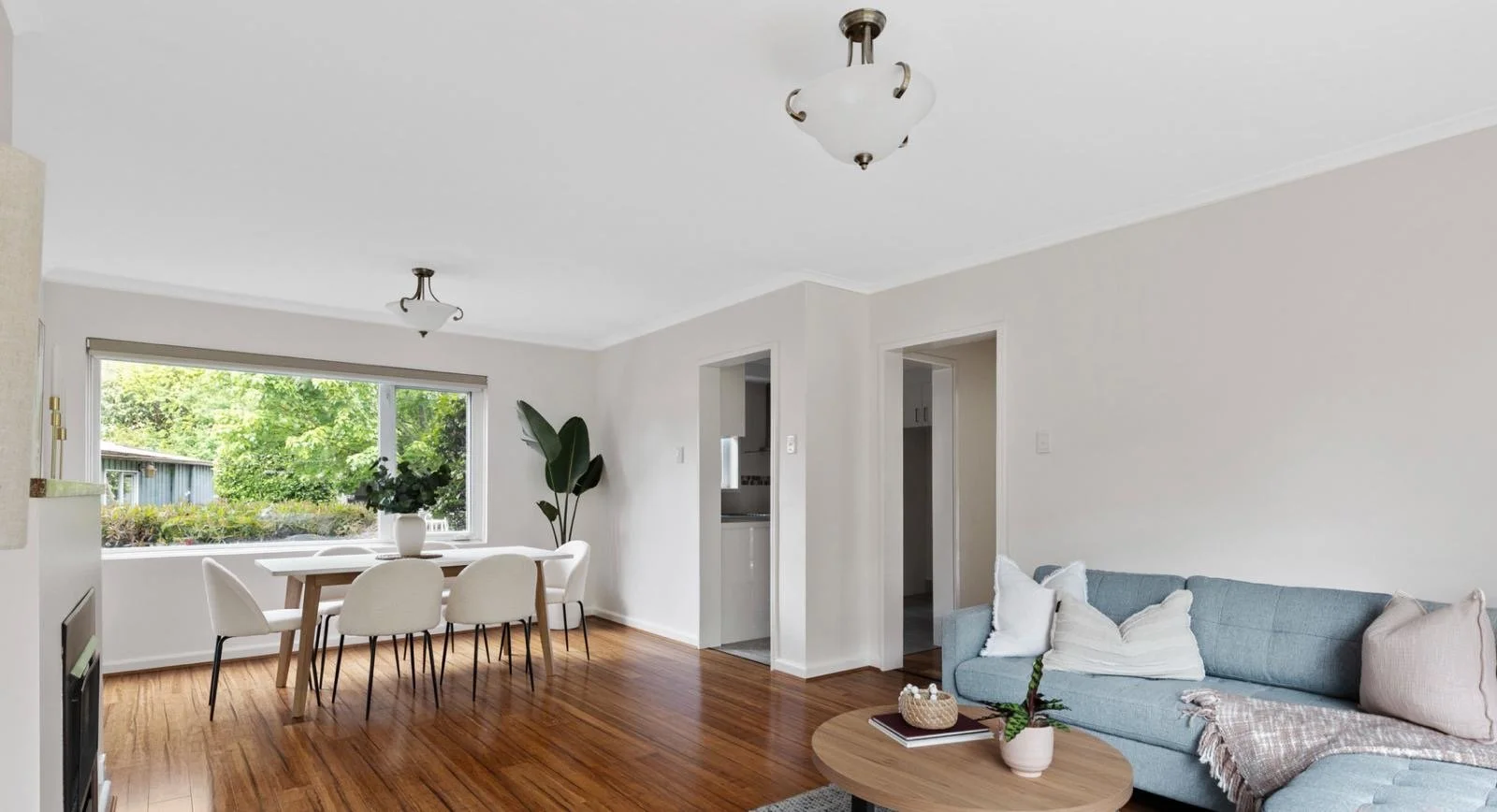 Living room with wooden floor, white walls, a large window with greenery outside, a light blue couch with pillows, a round wooden coffee table with a potted plant and books, a dining table with six white chairs, ceiling lights, and a potted plant nea