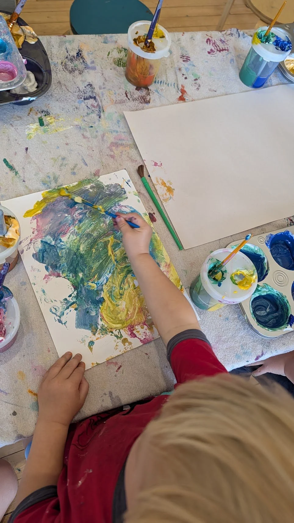 Child painting with colorful paints and brushes on paper, surrounded by paint containers and a paint palette on a table with a paint-splattered cloth.