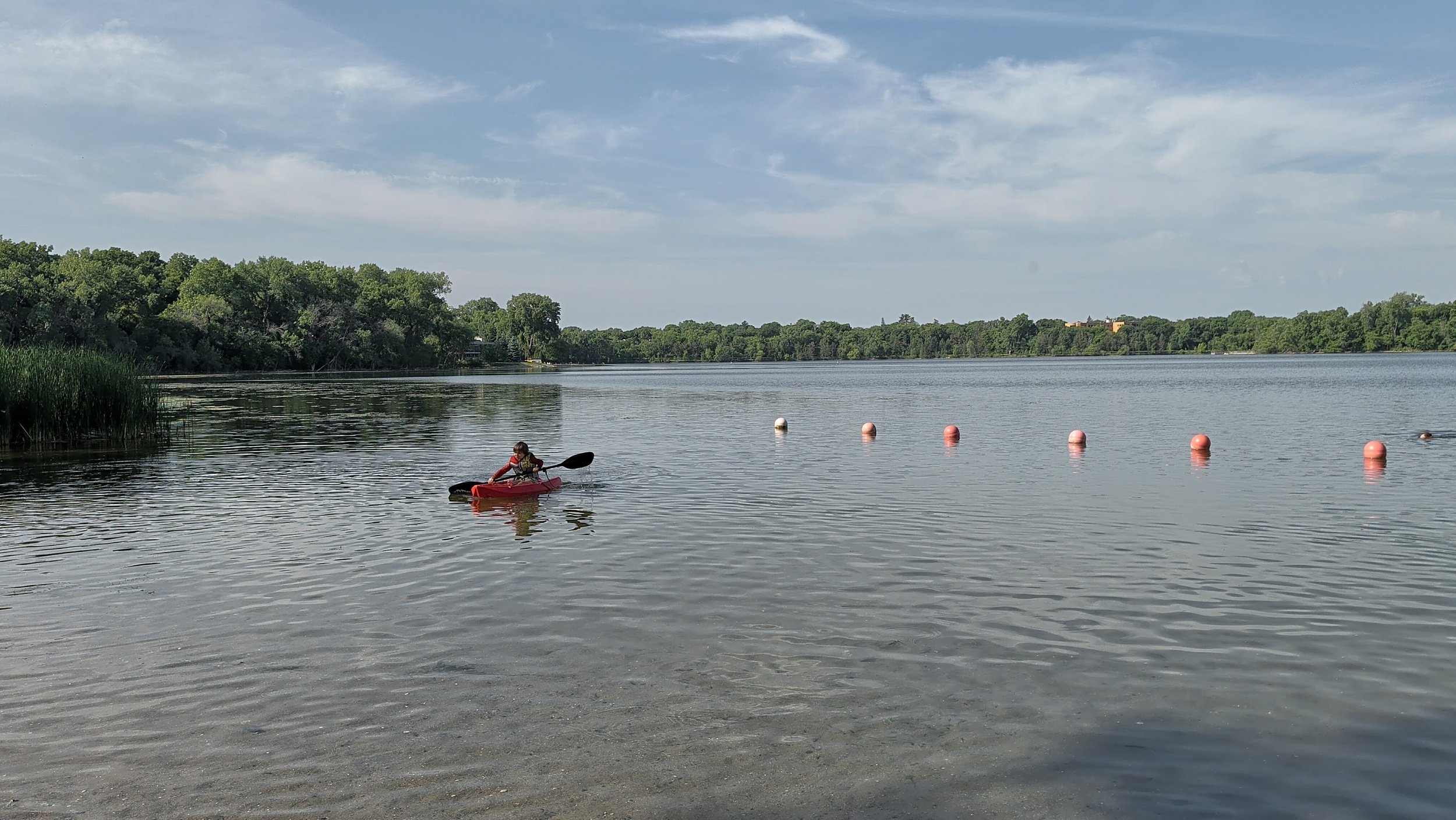 A person kayaking on a calm lake near a sandy shore with green trees in the background under a partly cloudy sky.