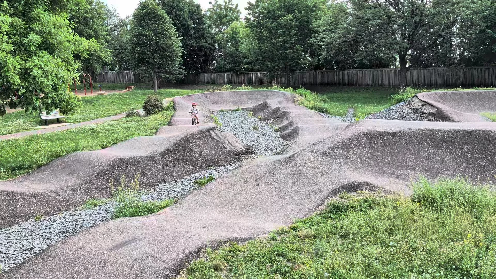 A small child riding a bicycle on a dirt skate park with concrete ramps and a gravel stream, surrounded by green grass and trees, with a wooden fence in the background.