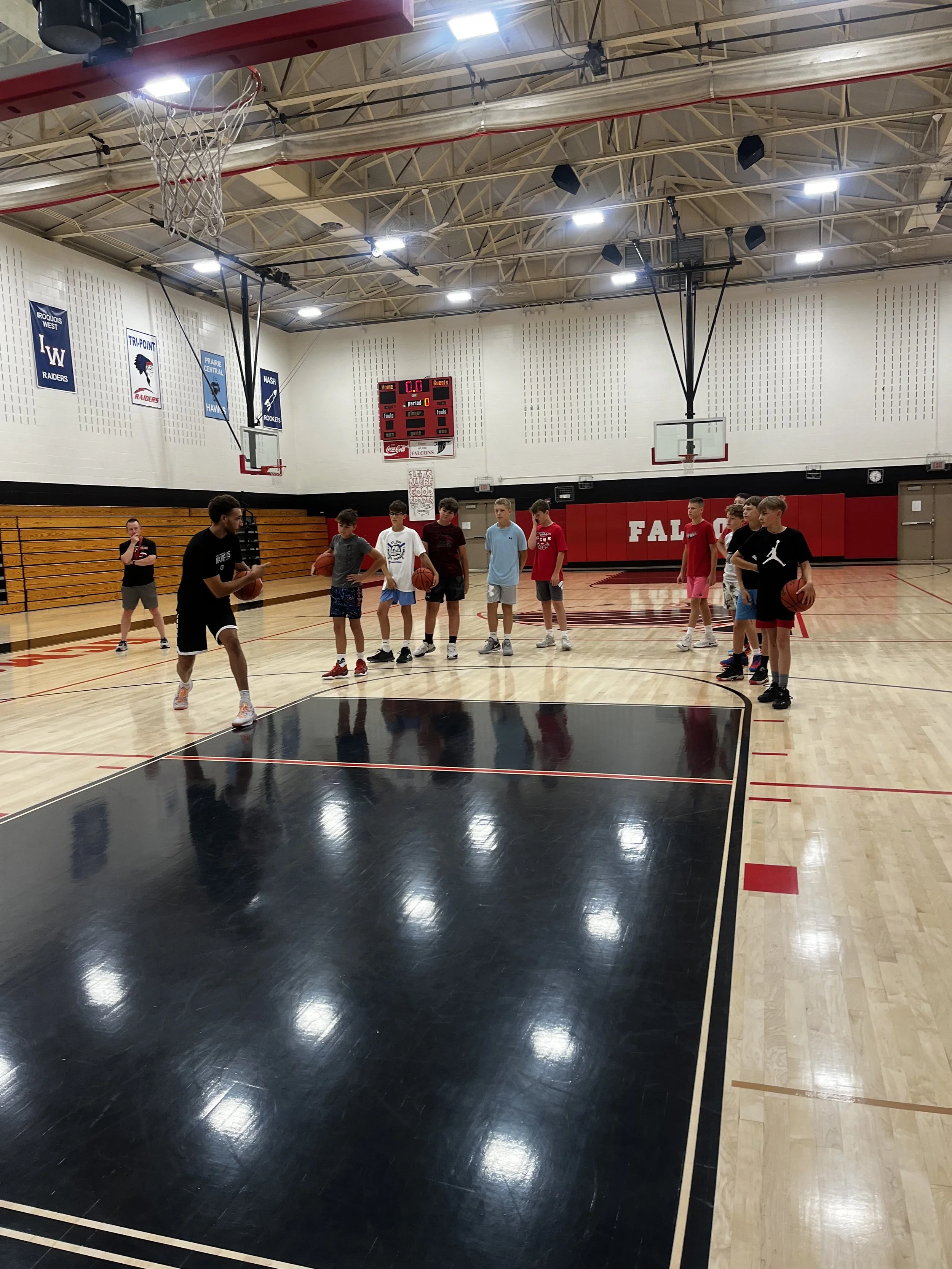 A gymnasium with children and a coach on a basketball court. The children are holding basketballs, and some are listening to instructions. The gym has banners on the wall, a scoreboard, and a basketball hoop.