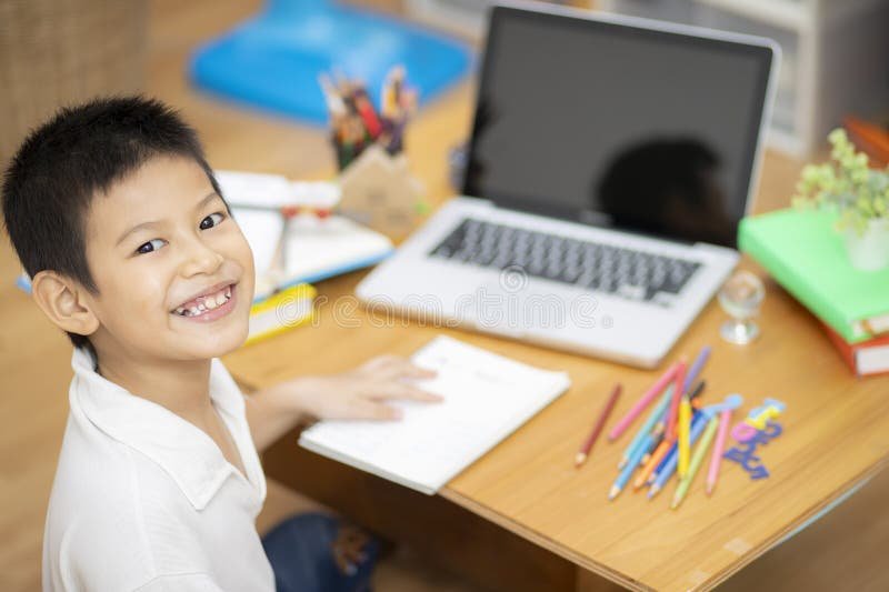 Smiling child at a desk with a laptop, notebook, and colorful pencils.