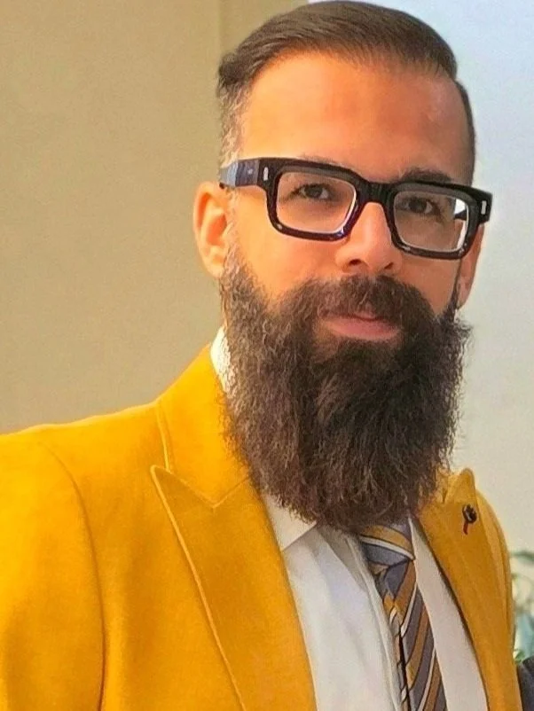 A man with glasses, a full beard, and a mustache wearing a yellow blazer, white shirt, and striped tie.