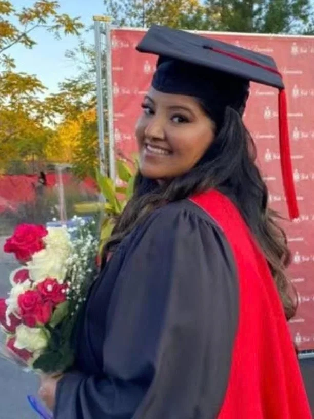 A young woman in a black graduation cap and gown holding a bouquet of red and white roses, smiling outdoors on graduation day.