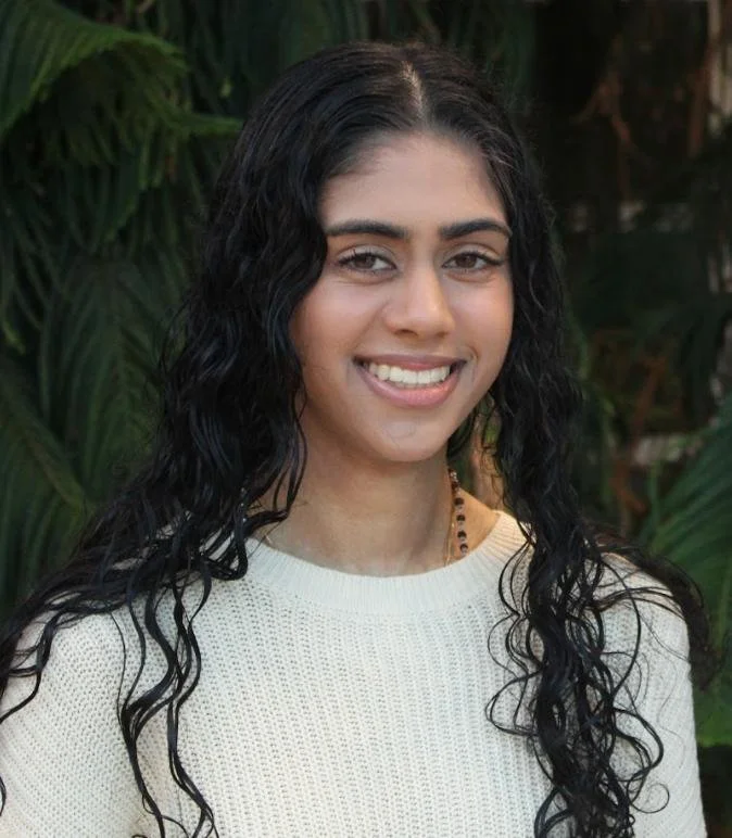 A young woman with curly black hair, smiling, wearing a white knit sweater and a beaded necklace, standing outdoors with green foliage in the background.