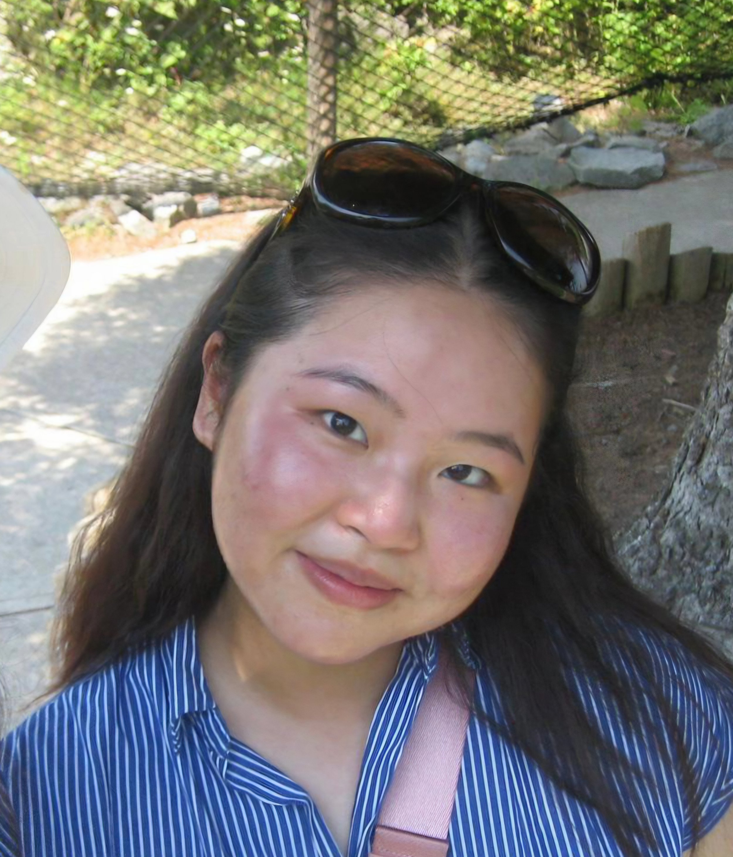 A woman with long dark hair wearing sunglasses on her head, a blue and white striped shirt, standing outdoors near a tree and a rocky area.