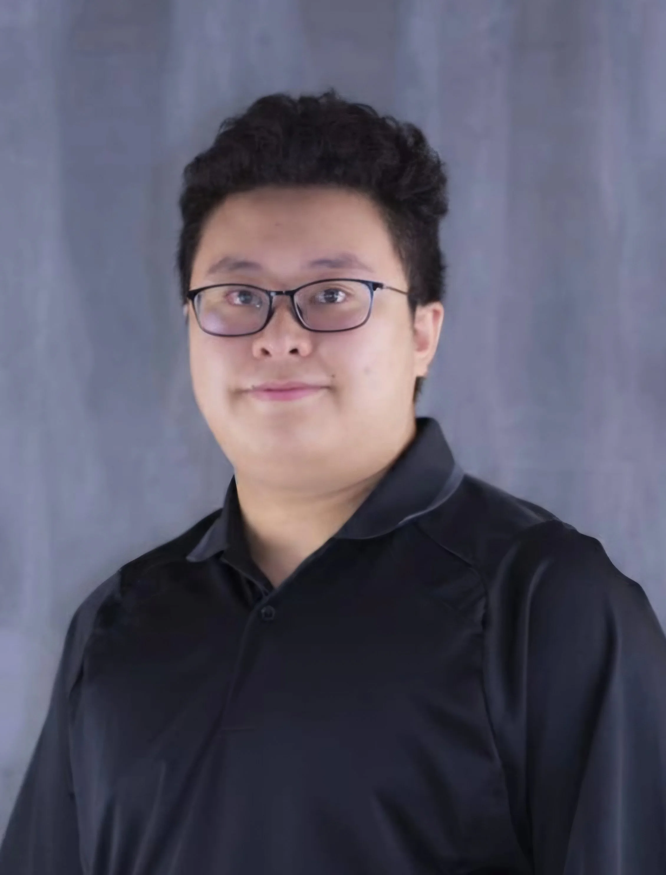 A young man with glasses and dark hair, wearing a black shirt, standing in front of a gray background.