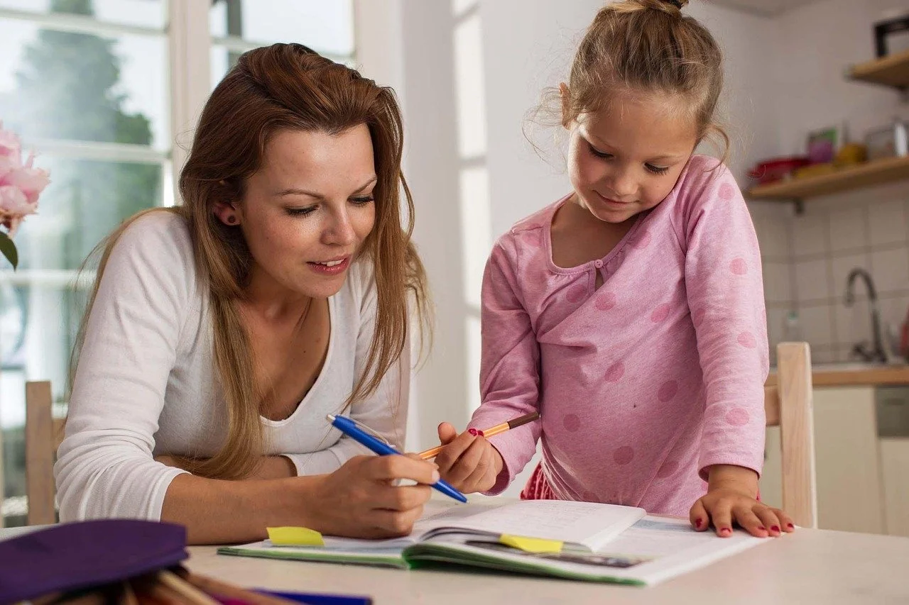A woman and a young girl sitting at a table, looking at an open book together, with the woman holding a blue pen and the girl holding a yellow pencil.