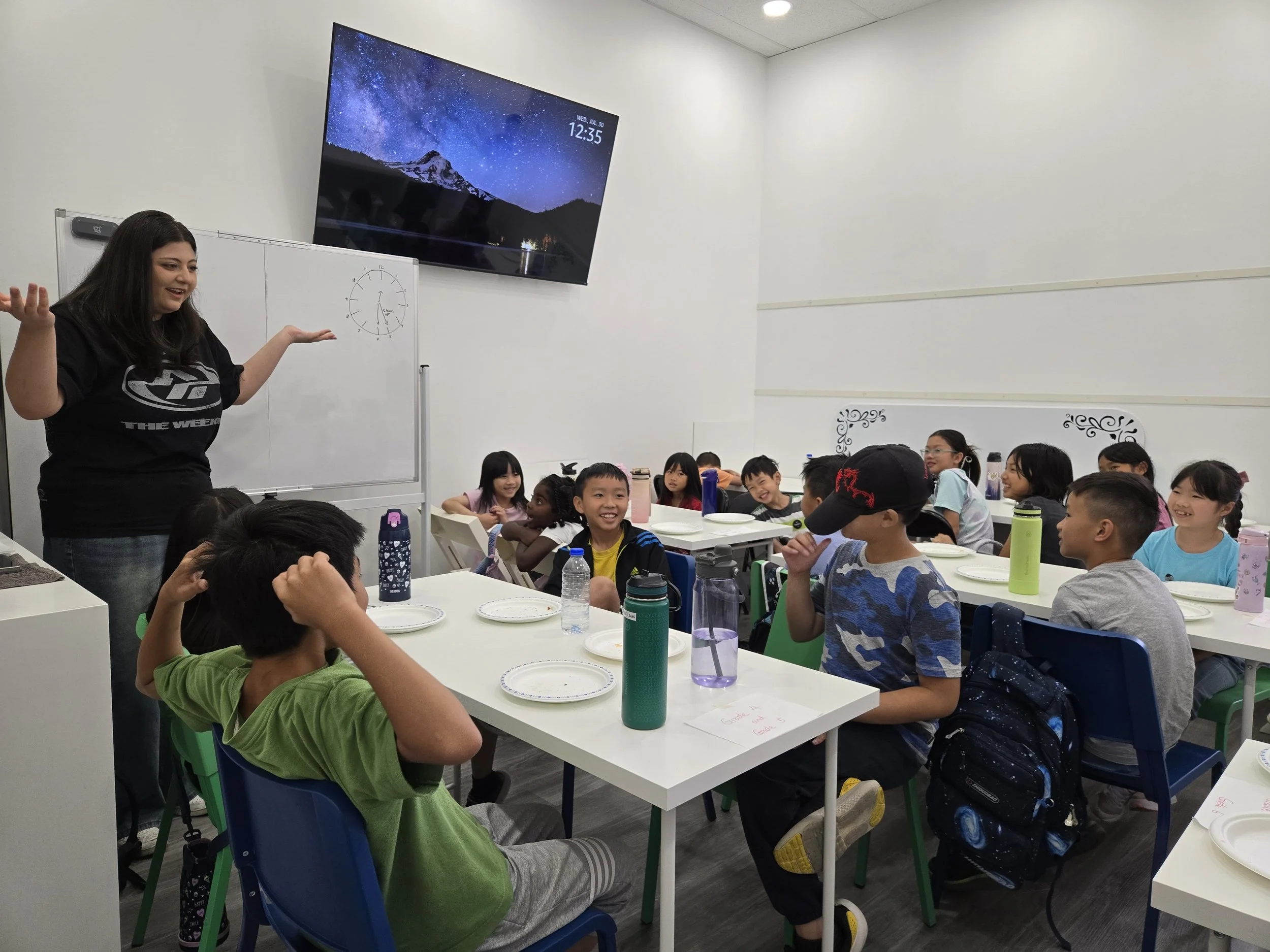A female teacher standing in front of a classroom of elementary school students, all smiling and engaging with her, with a whiteboard and a large digital screen showing a starry night sky and mountain landscape in the background.