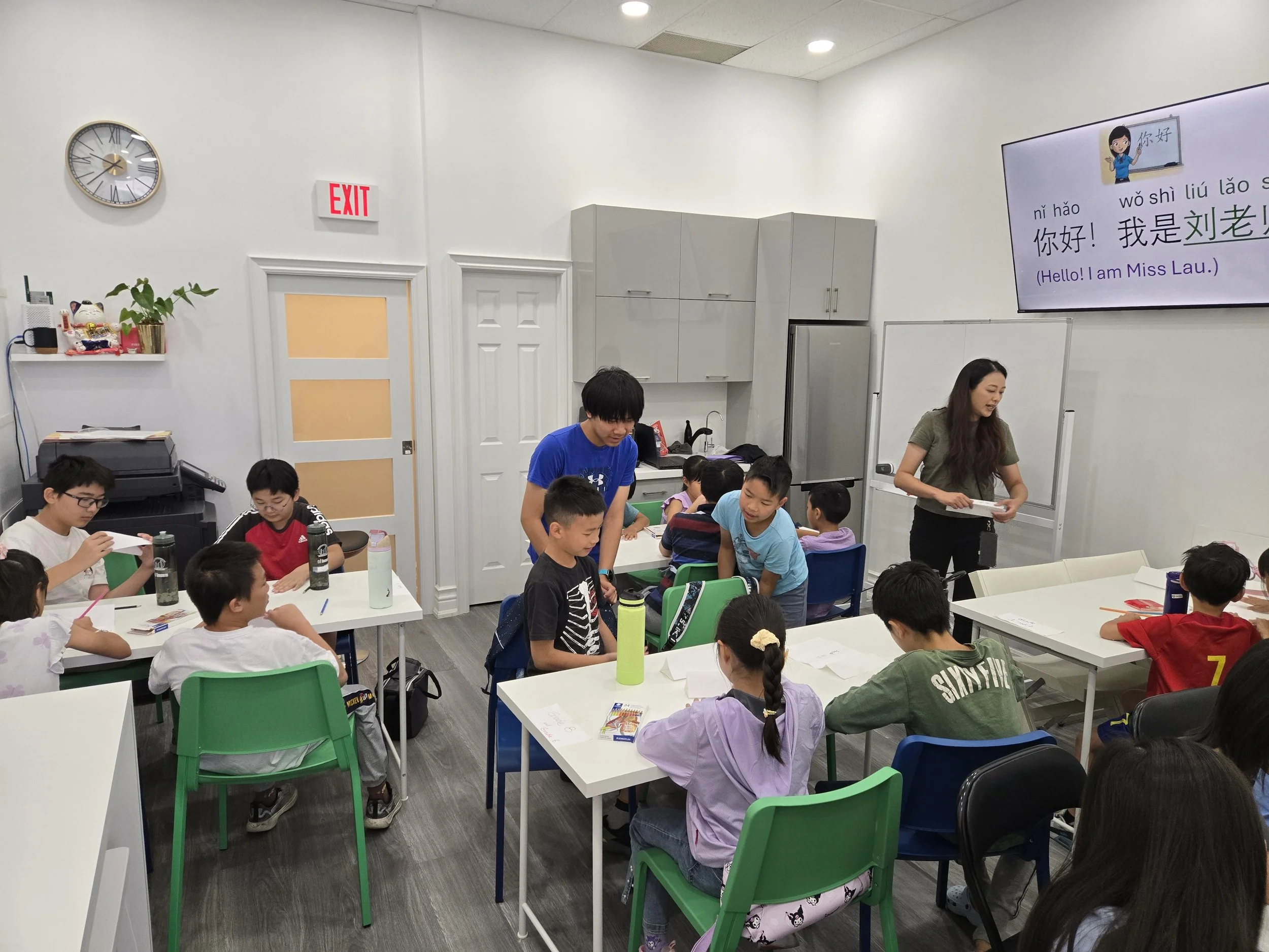 A classroom with children seated at desks, a teacher standing near a whiteboard, and a large screen displaying Chinese characters and English translations for a language lesson.