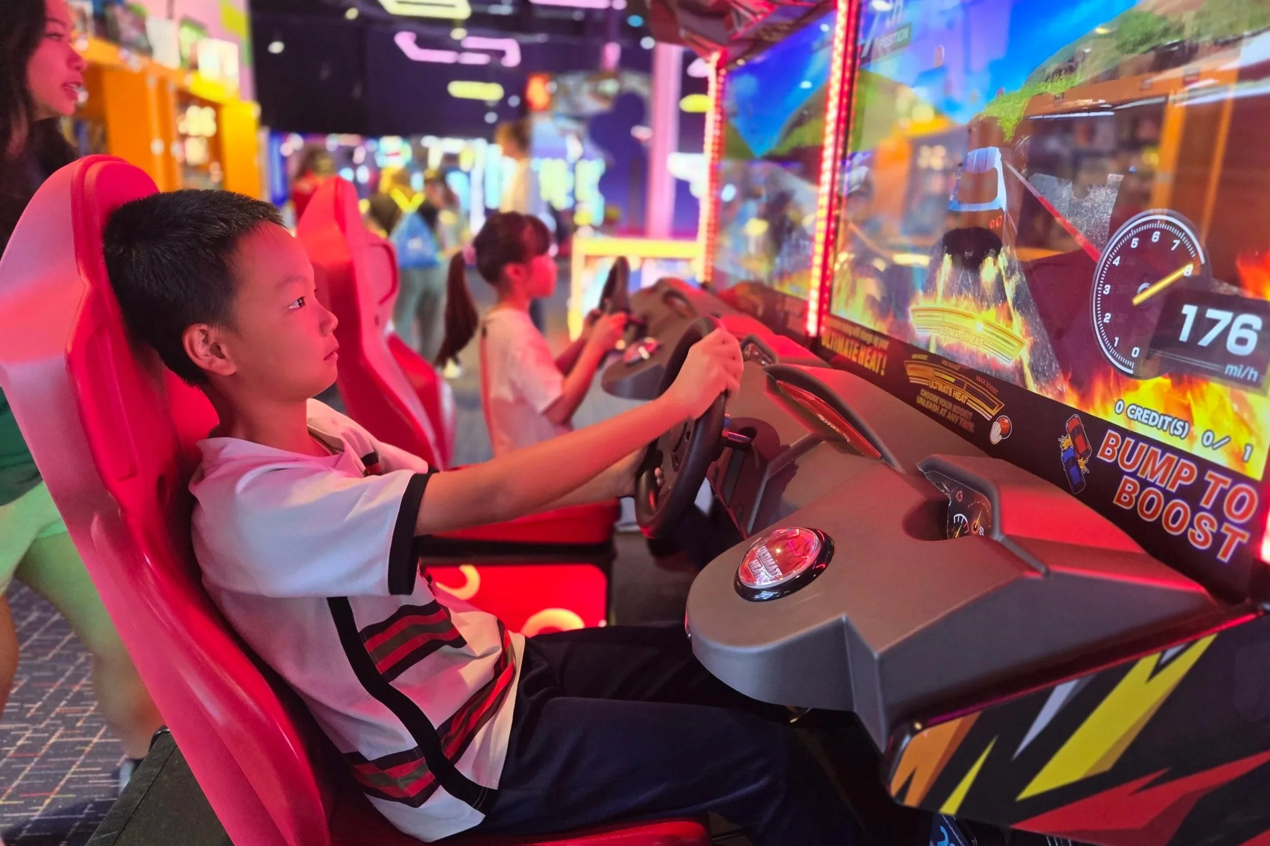 A young boy with short black hair seated in a red racing arcade game seat, holding a steering wheel, playing a racing game with bright, colorful graphics on the screen. Other children are visible in the background also playing arcade games in a vibrant arcade setting.