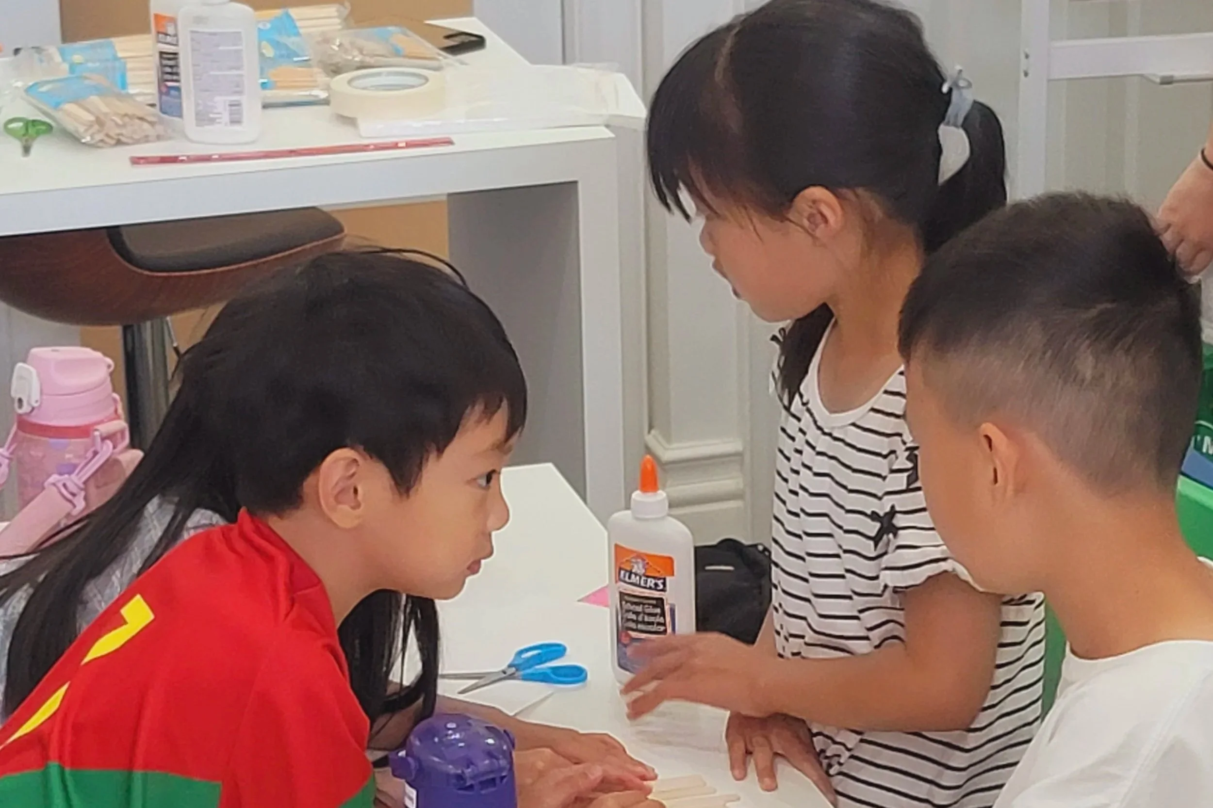 Three children around a table engaging in a craft activity, with supplies like scissors, glue, and water bottles visible on the table.