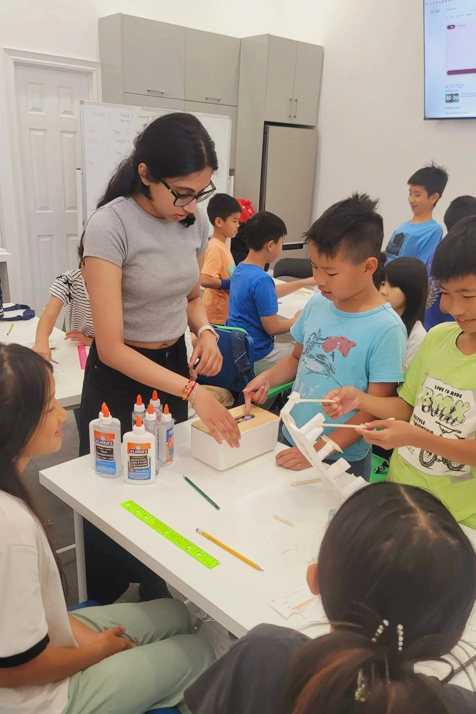 A teacher at a classroom table assisting children with a craft project involving glue, wood sticks, and paper, with children smiling and engaging in the activity