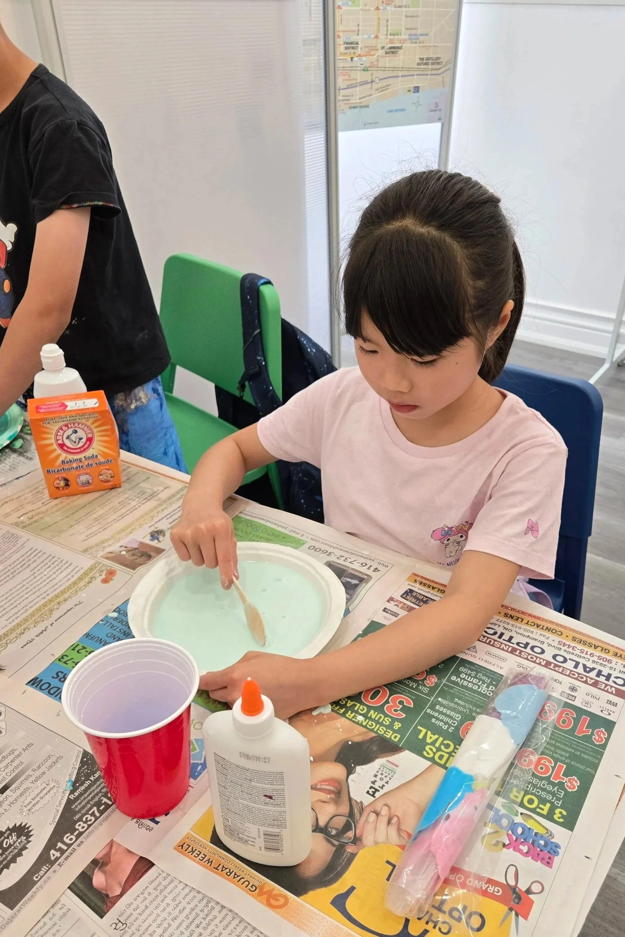 A young girl sitting at a table with a bowl of a blue liquid, a red plastic cup, and craft supplies, mixing something with a wooden stick, in what appears to be a classroom or craft area.